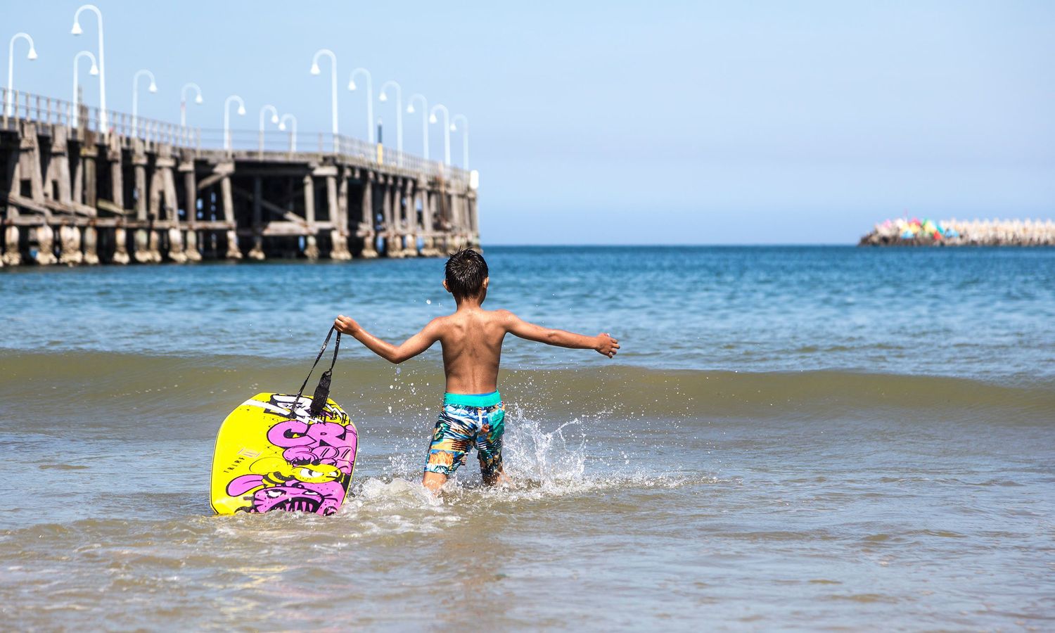 Colour image of a young boy running into the surf with a boogie board, full of vitality and summer energy. Photo by Raymond Mather.