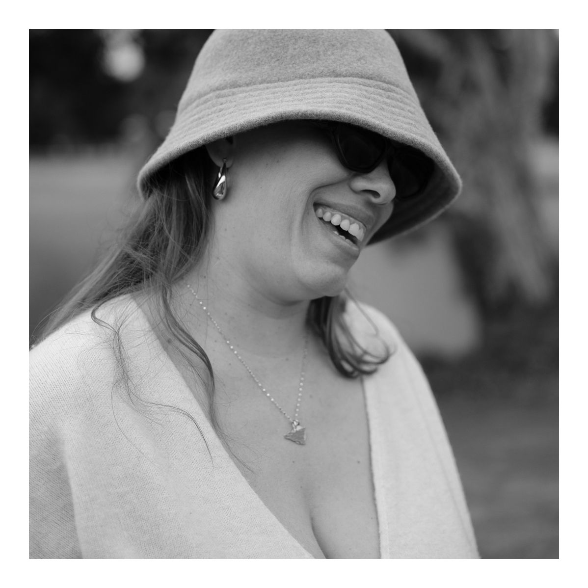 Black and white candid outdoor photo of mother smiling beneath a soft bucket hat. Photo by Raymond Mather.