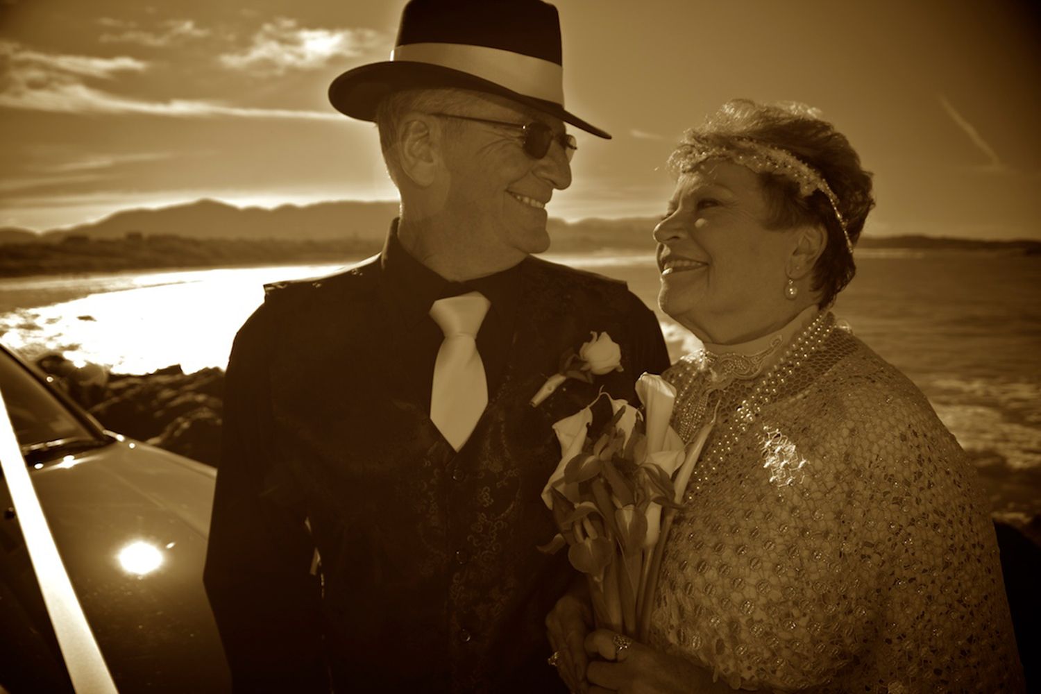 Sepia-toned portrait of couple in vintage wedding attire, standing by the sea at sunset. Photo by Raymond Mather.