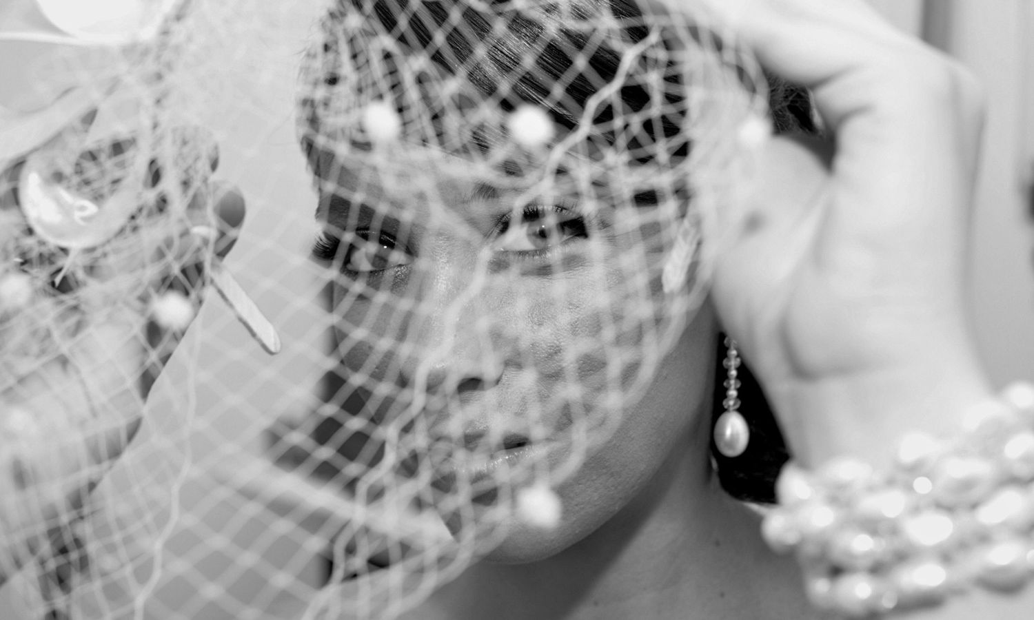 Monochrome close-up of bride adjusting her birdcage veil, evoking timeless grace and quiet reflection. Photo by Raymond Mather.