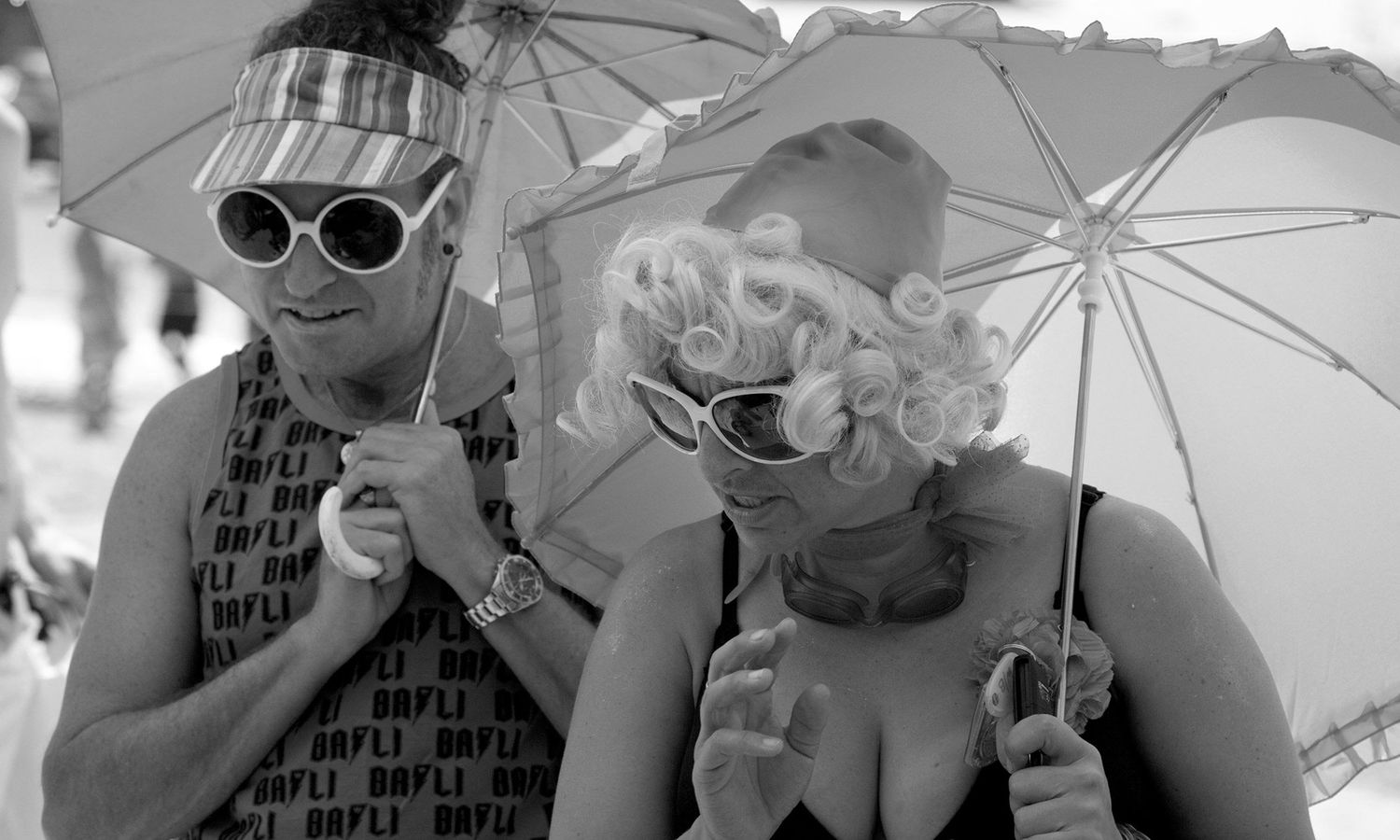 Monochrome street photo of friends dressed playfully in Drag with umbrellas at an outdoor festival. Photo by Raymond Mather.