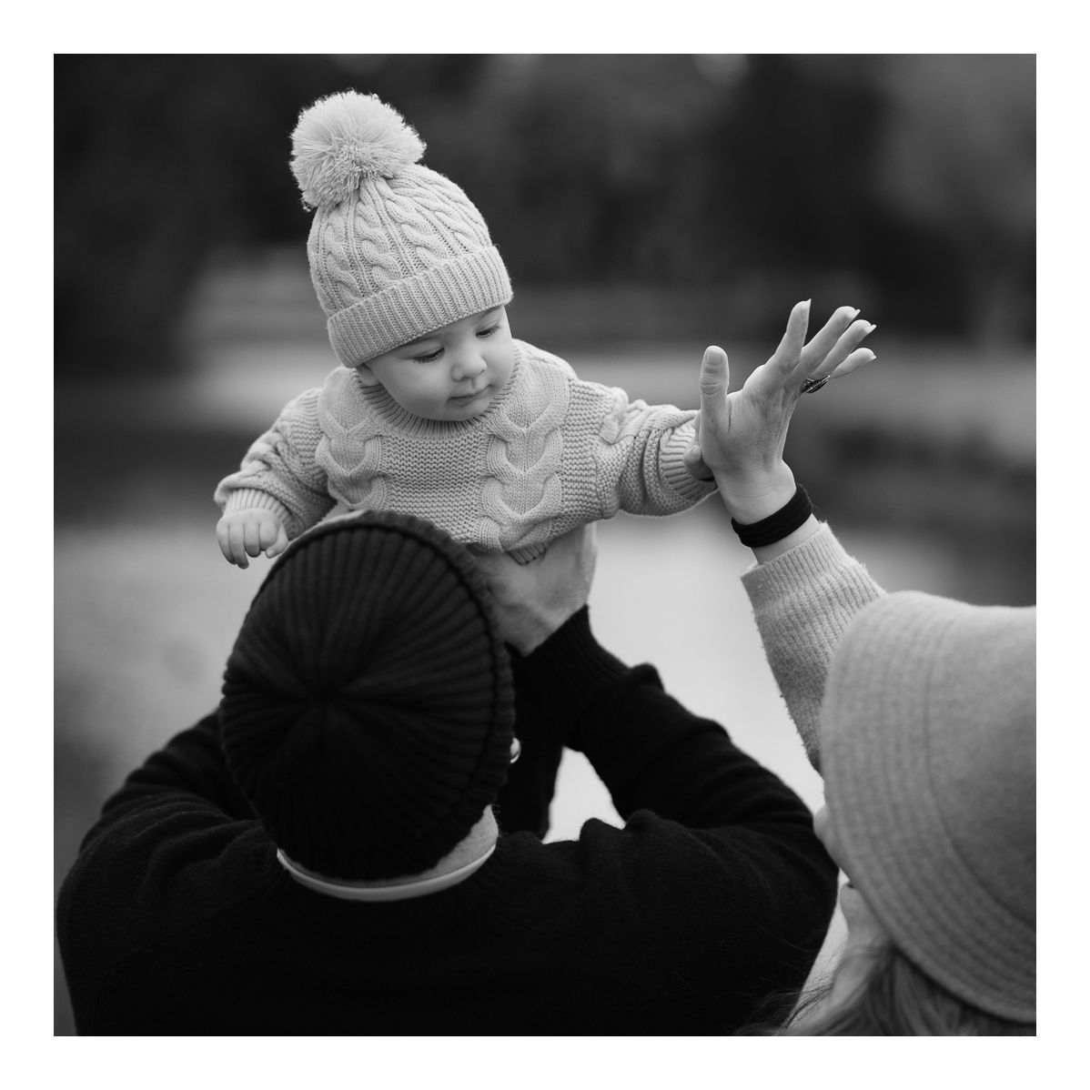Black and white candid outdoor photo of parents lifting their baby playfully in the park. Photo by Raymond Mather.