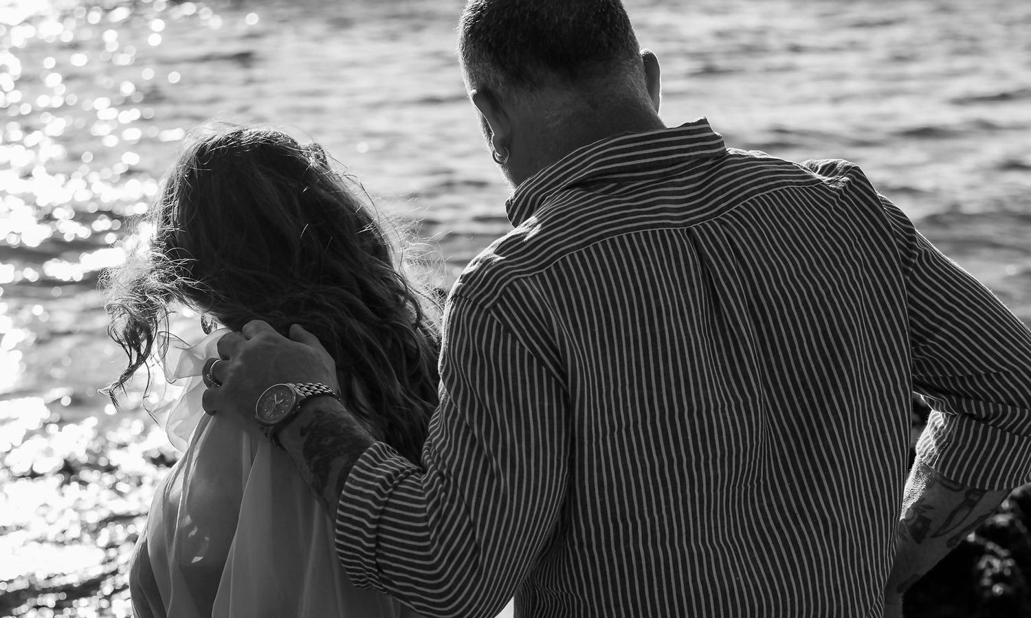 Monochrome candid beach photo of expectant couple walking together beside the water. Photo by Raymond Mather.
