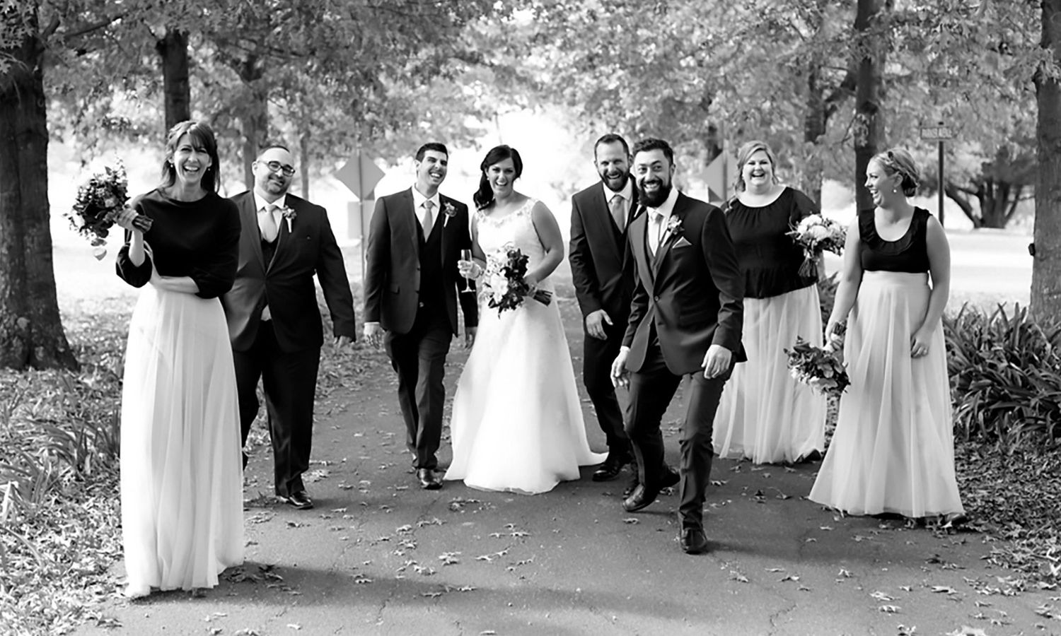 Black and white version of bridal party walking along autumn path, highlighting natural light, classic composition. Photo by Raymond Mather.