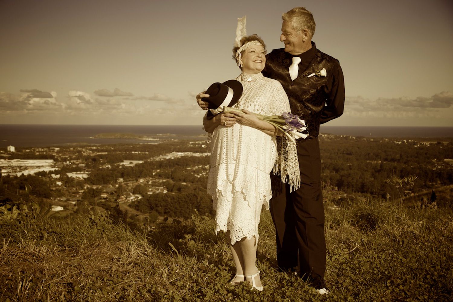 Sepia-toned wedding portrait of couple in vintage attire standing on a hilltop at sunset. Photo by Raymond Mather.
