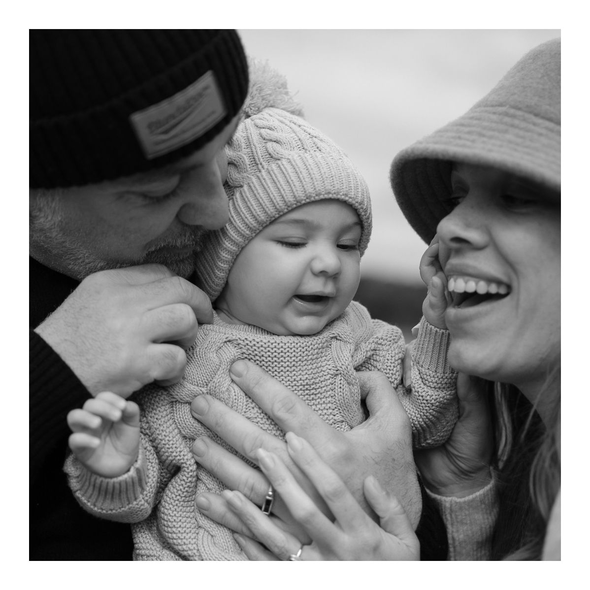 Black and white candid outdoor photo of parents laughing with baby in cosy winter outfits. Photo by Raymond Mather.