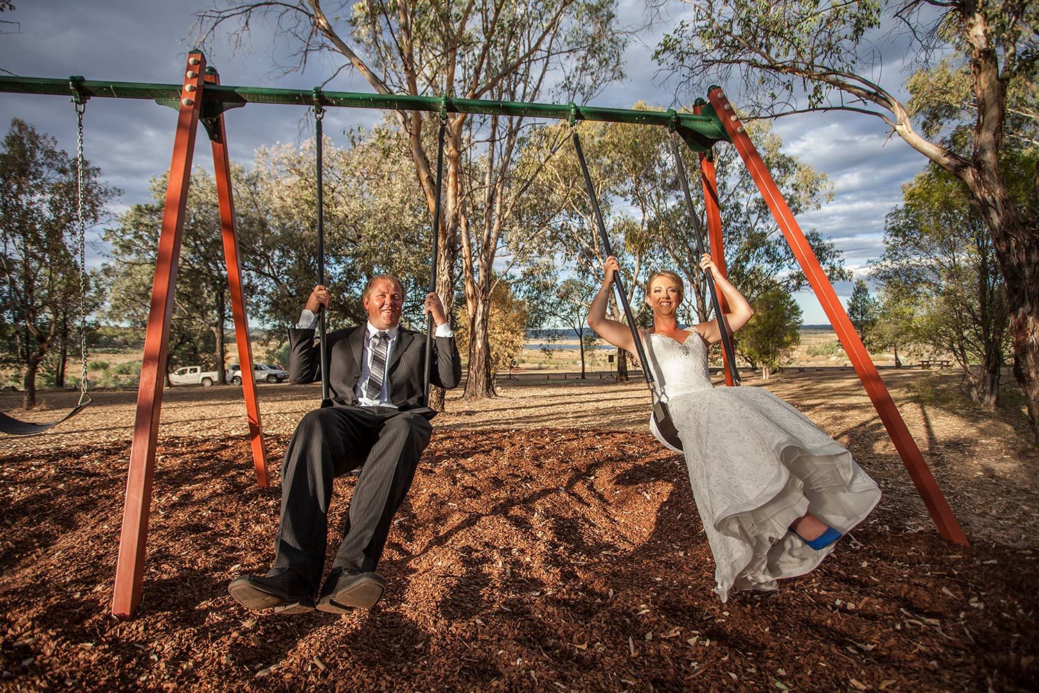 Colour wedding photo of bride and groom joyfully swinging together beneath clear afternoon skies. Photo by Raymond Mather.