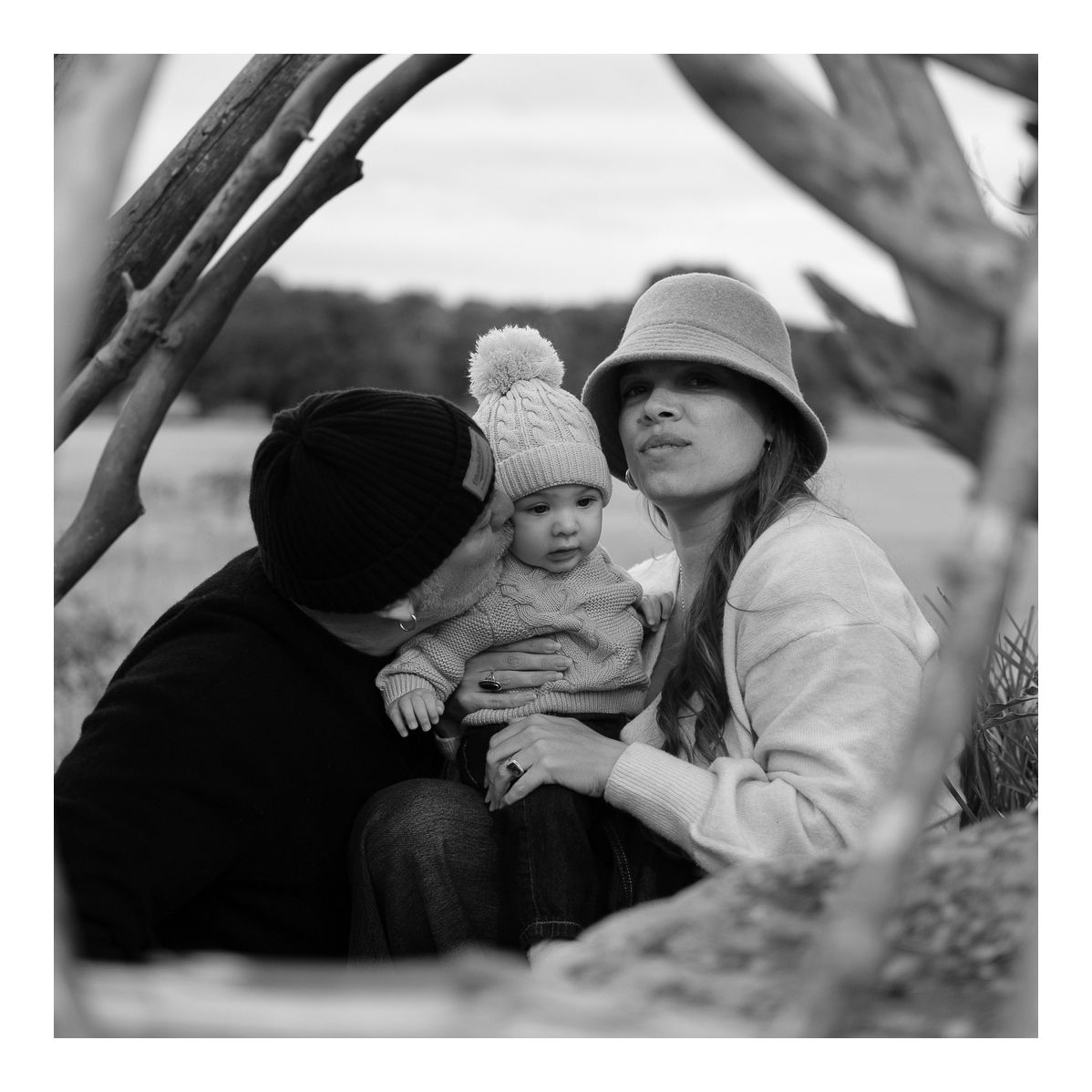 Black and white outdoor portrait of family sitting beneath driftwood shelter with baby. Photo by Raymond Mather.