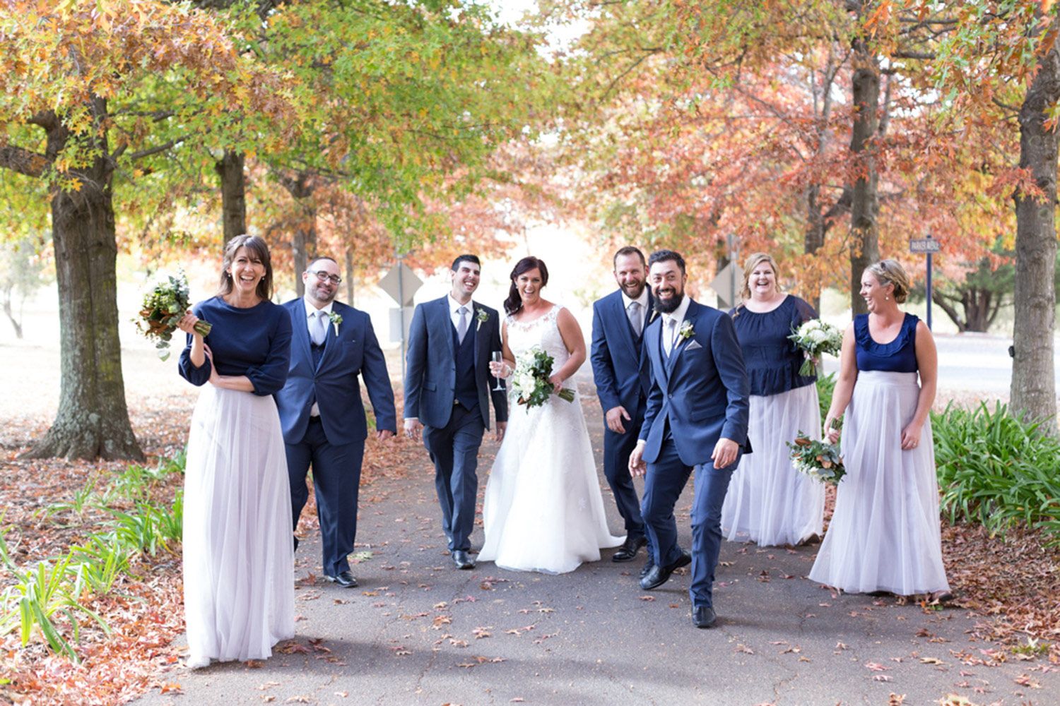 Colour bridal party photo with bride, groom, and attendants walking together along a tree-lined path. Photo by Raymond Mather.