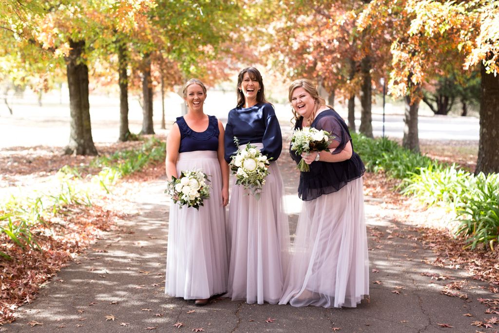 Colour portrait of bridesmaids walking through autumn trees, holding bouquets and smiling in soft morning light. Photo by Raymond Mather.