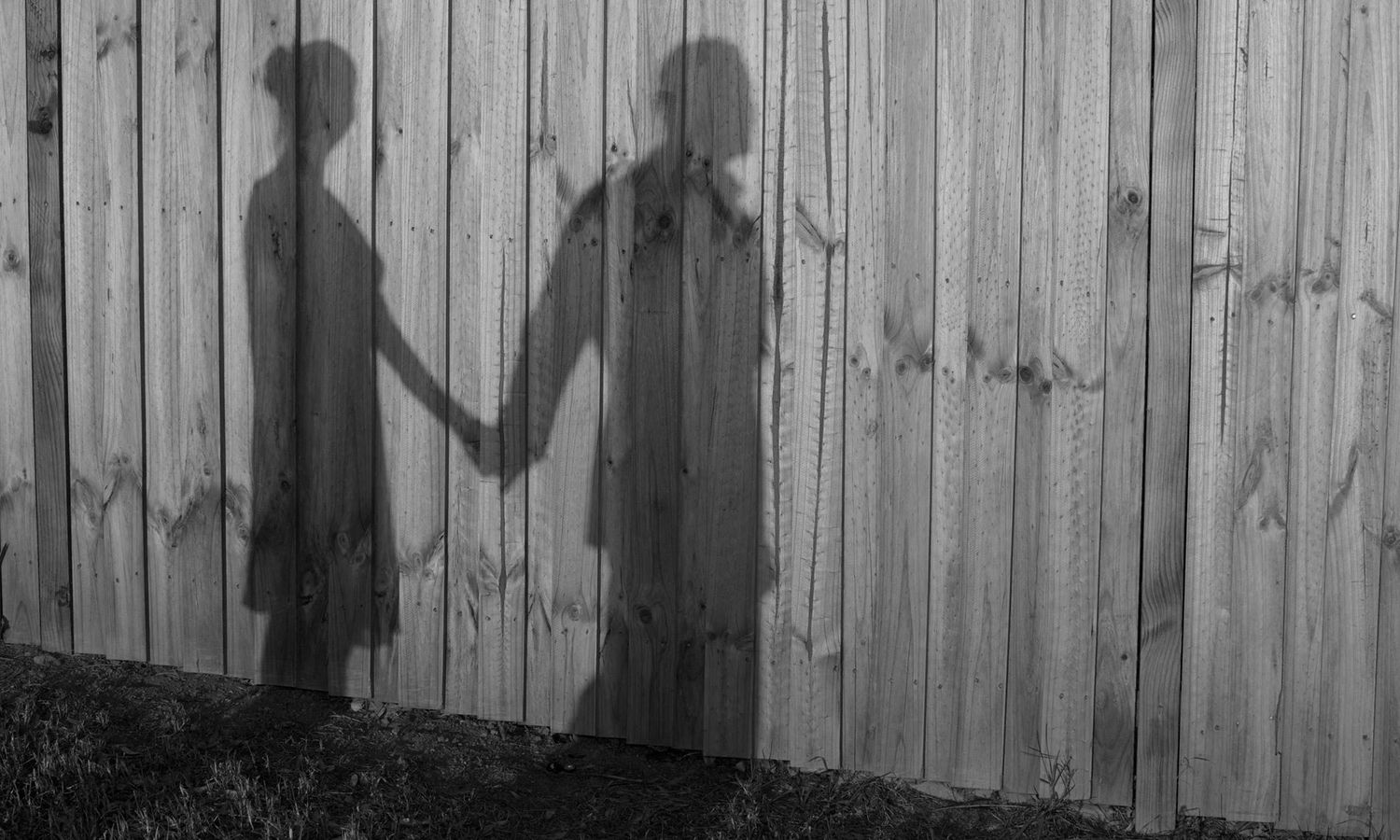 Black and white artistic image of couple’s silhouettes holding hands against a fence. Photo by Raymond Mather.