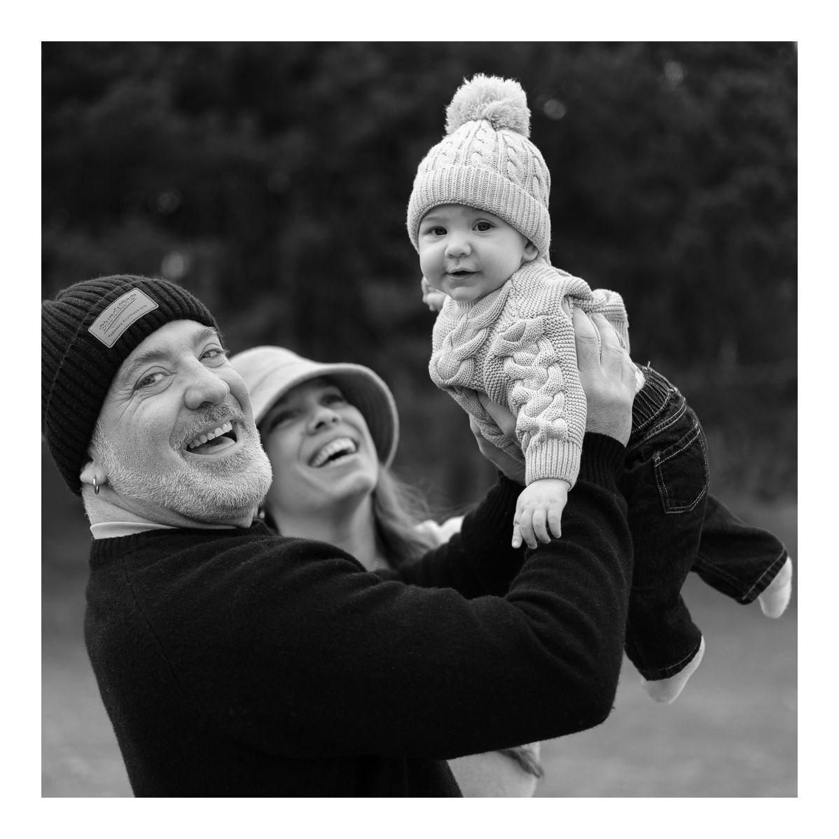 Monochrome candid outdoor photo of parents lifting smiling baby into the air. Photo by Raymond Mather.