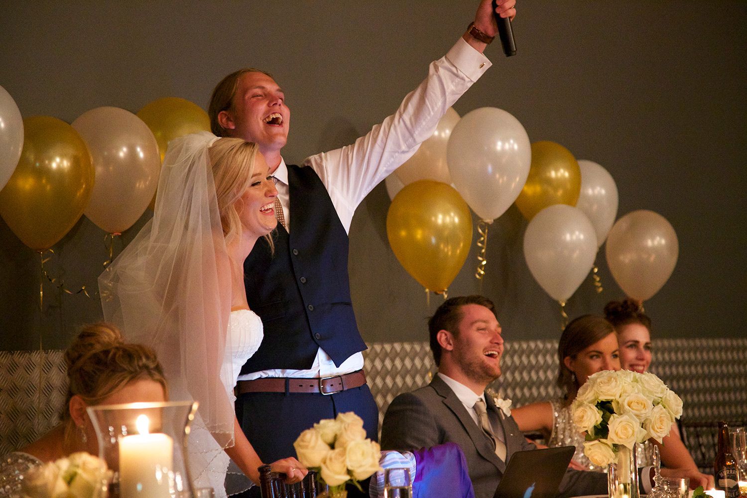 Colour reception image of newlyweds celebrating at their wedding dinner surrounded by friends and golden balloons. Photo by Raymond Mather.