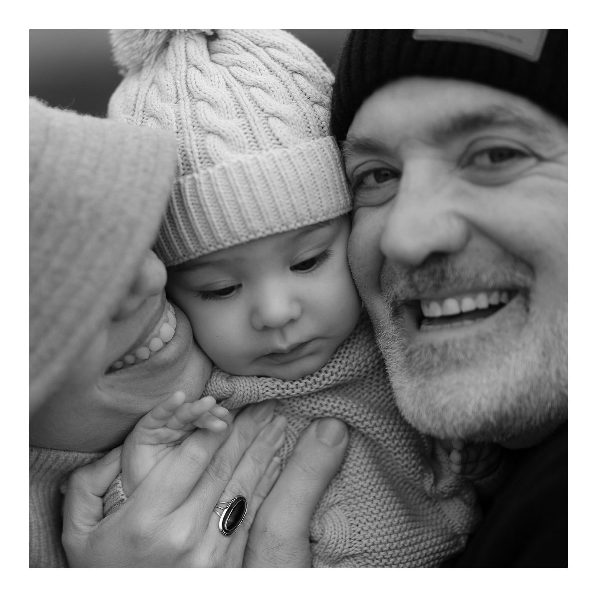 Monochrome candid portrait of mother kissing baby’s cheek as father smiles warmly. Photo by Raymond Mather.