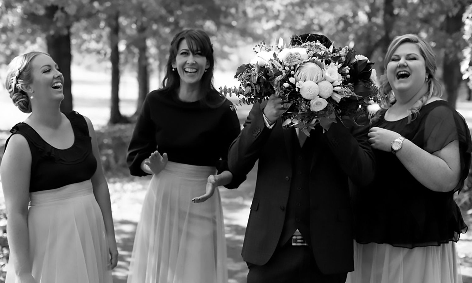 A group of wedding party members laugh together outdoors in formal attire. Photography by Raymond Mather.