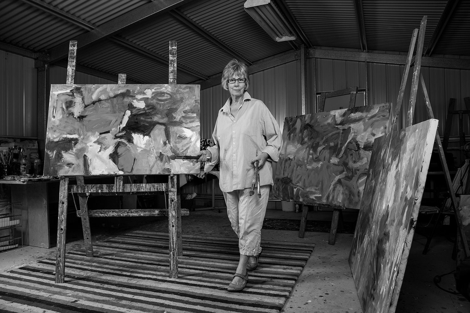 Black and white candid photo of artist standing in studio surrounded by abstract canvases. Photo by Raymond Mather.