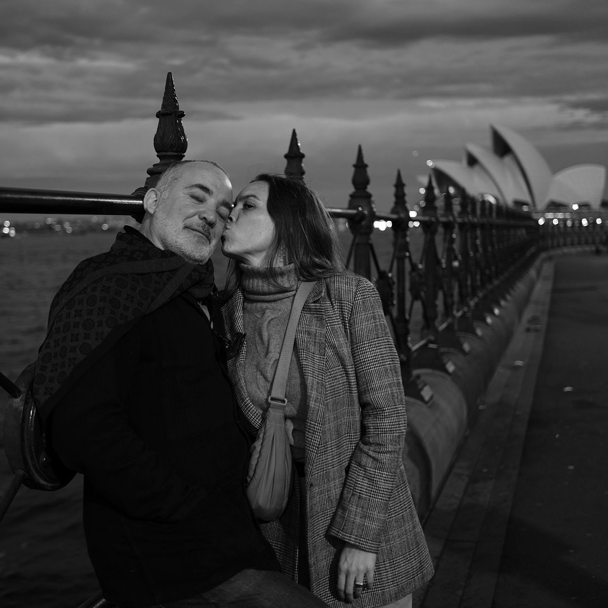 Black and white candid photo of couple kissing on Sydney Harbour walkway at dusk. Photo by Raymond Mather.