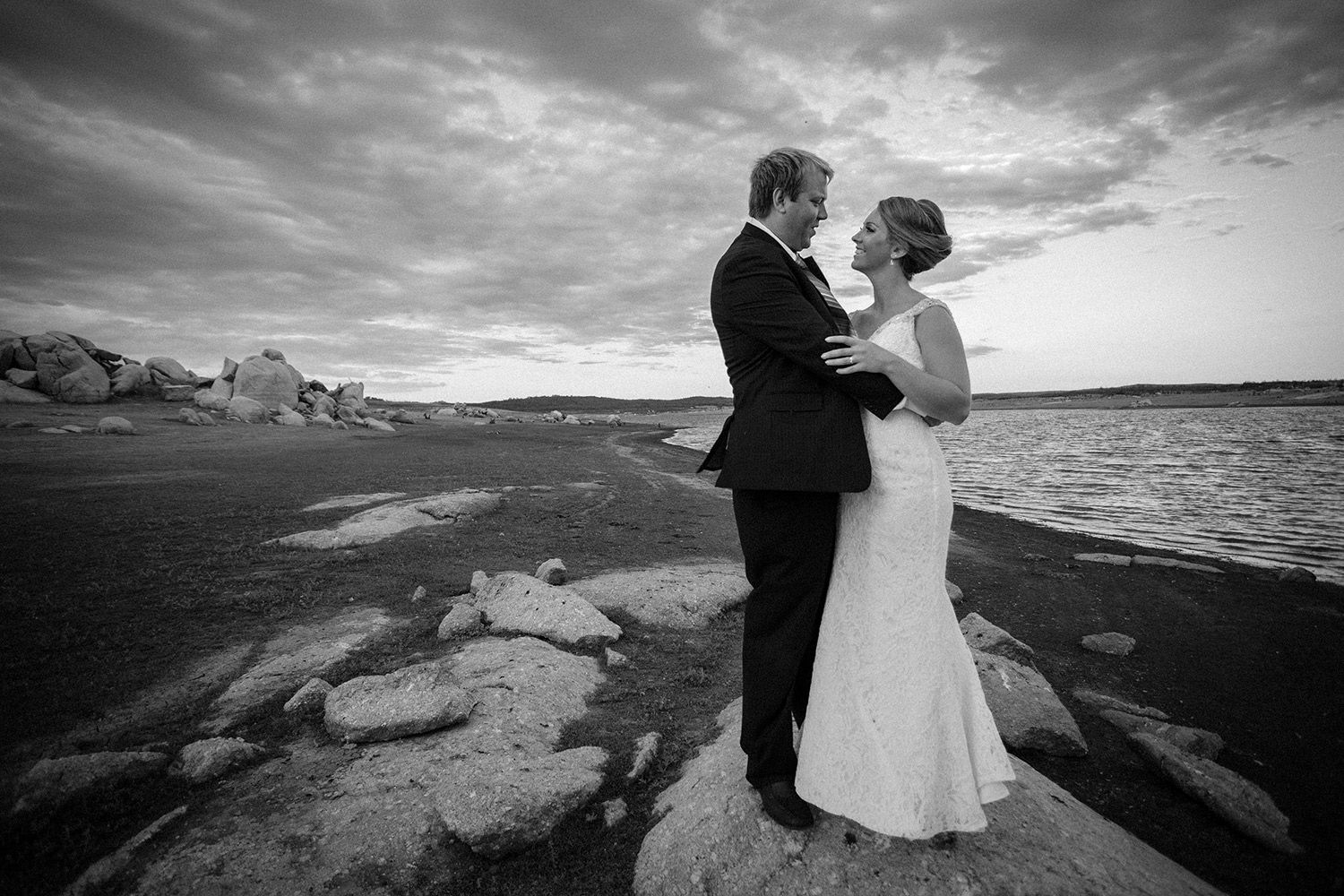 Black and white portrait of bride and groom embracing beside the lake under dramatic skies. Photo by Raymond Mather.
