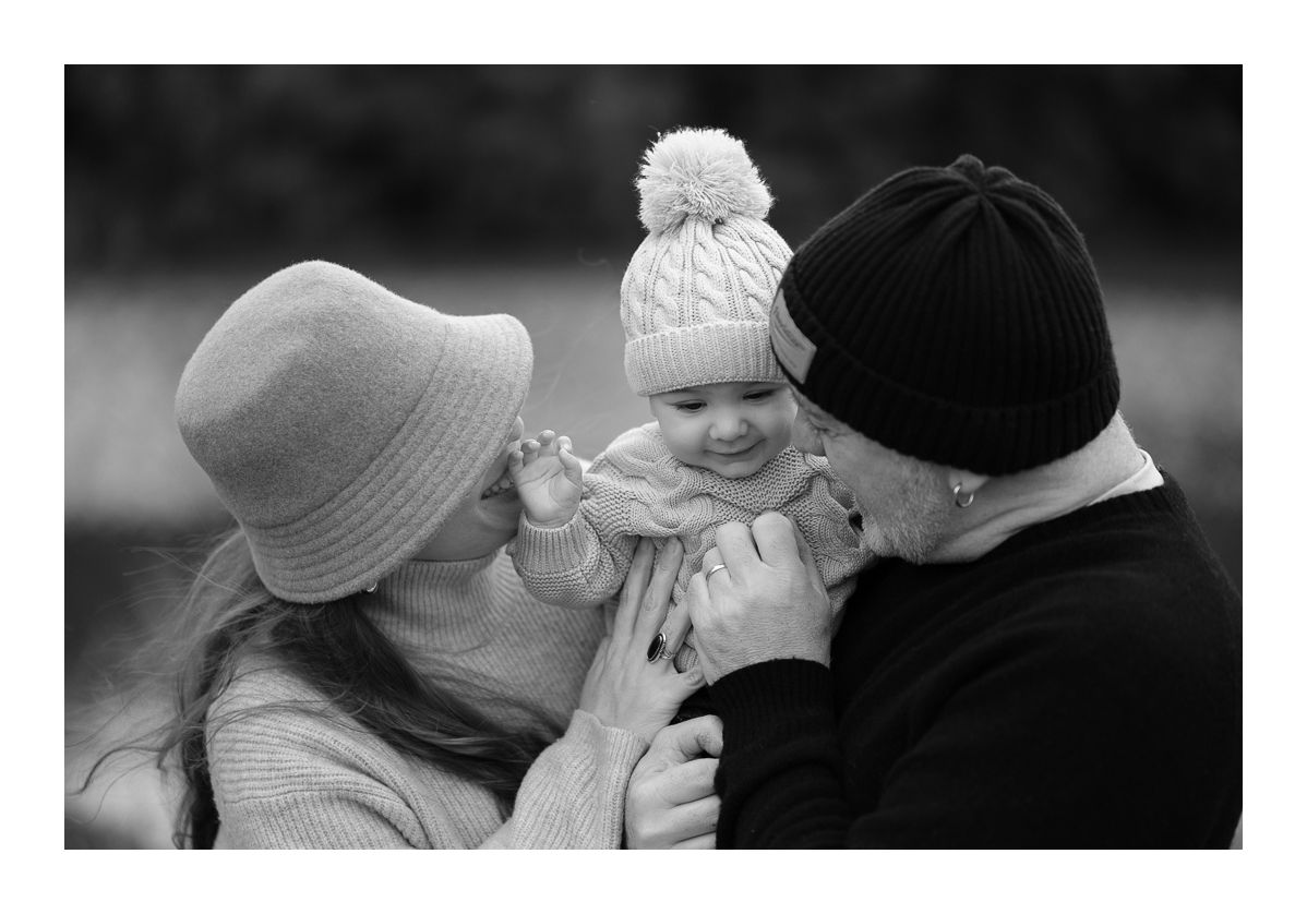Monochrome outdoor family portrait of parents kissing baby while sharing laughter. Photo by Raymond Mather.