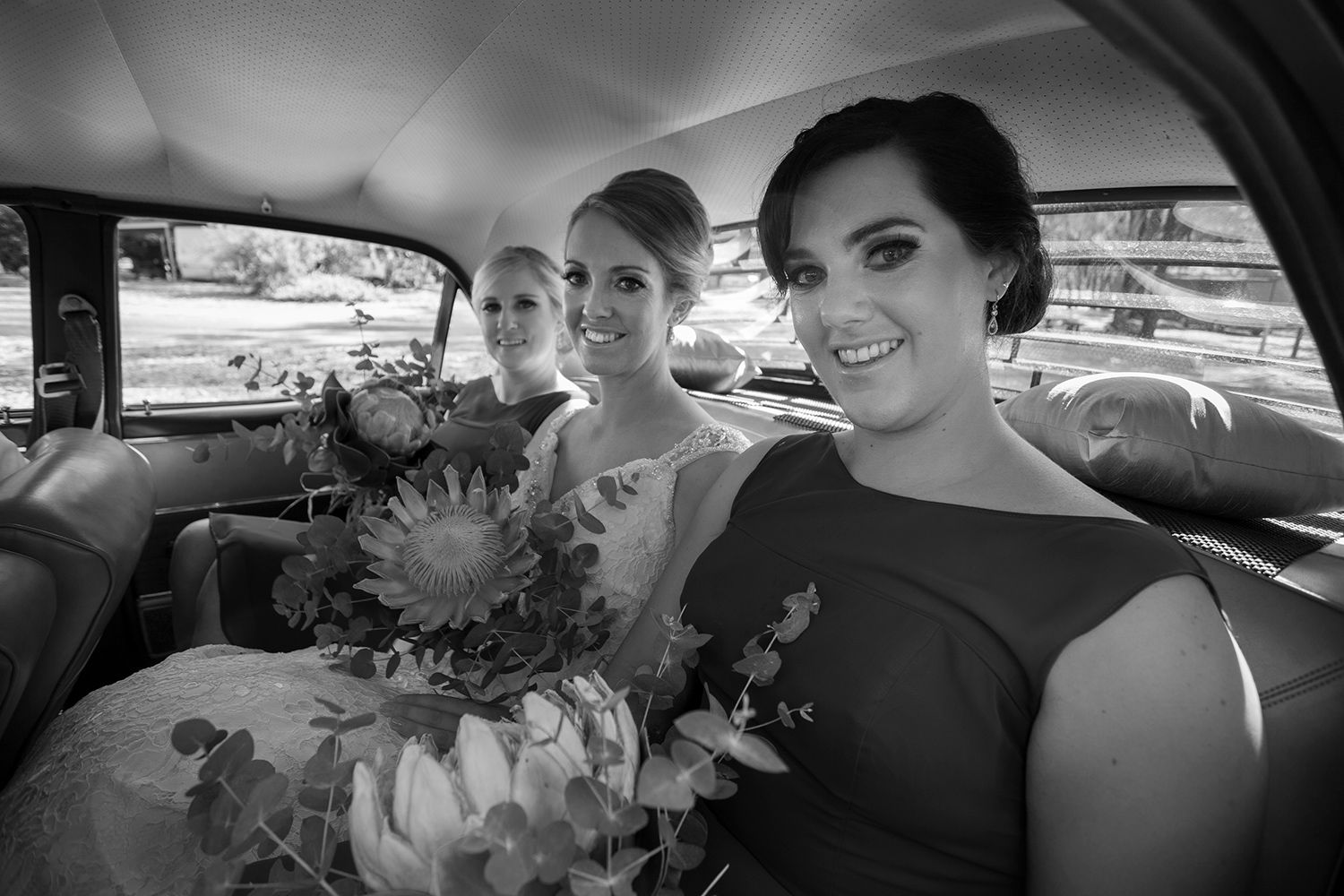 Monochrome photo of bridesmaids smiling from the backseat of a vintage car, framed by soft natural light. Photo by Raymond Mather.
