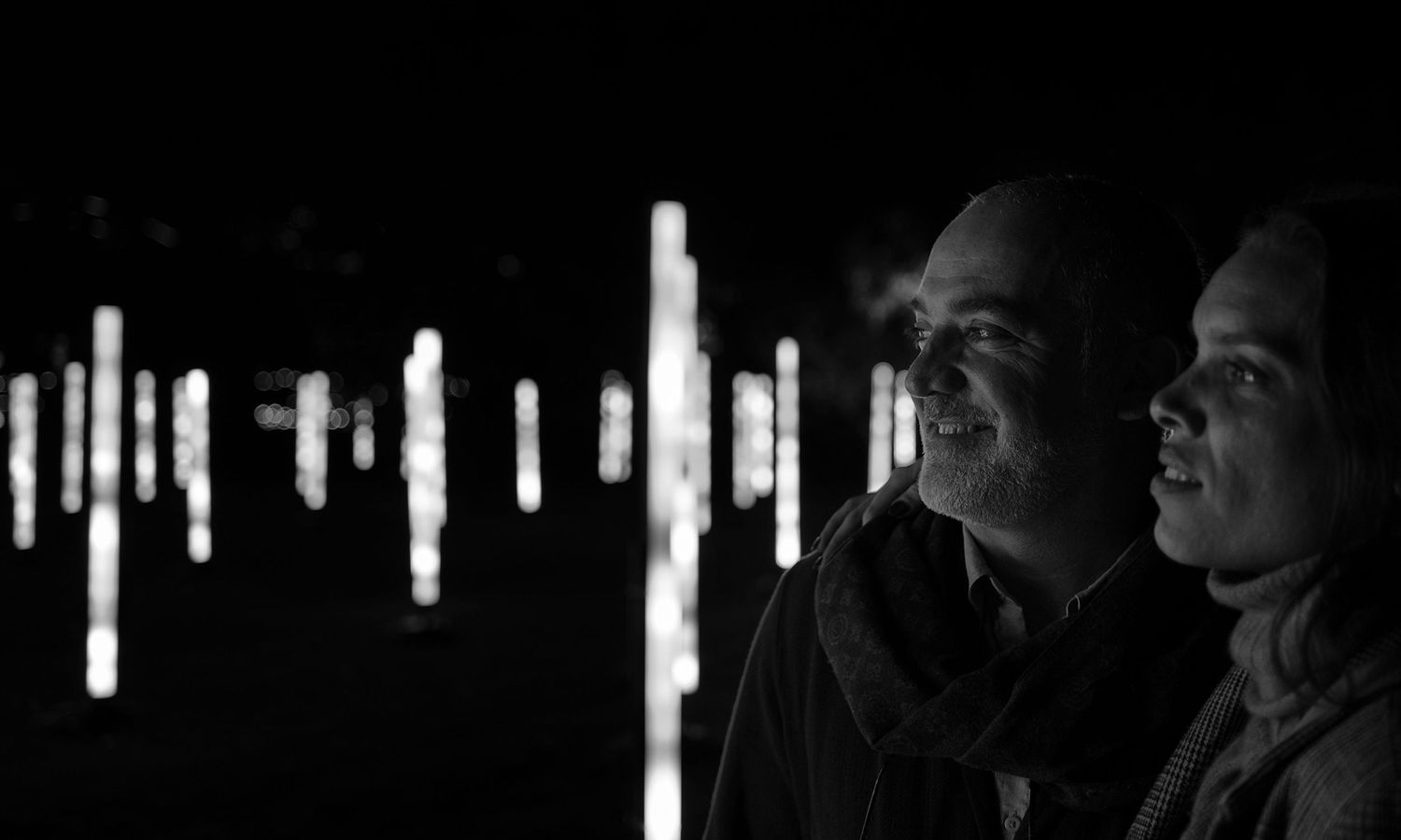 Monochrome night photo of couple standing together among glowing light columns. Photo by Raymond Mather.