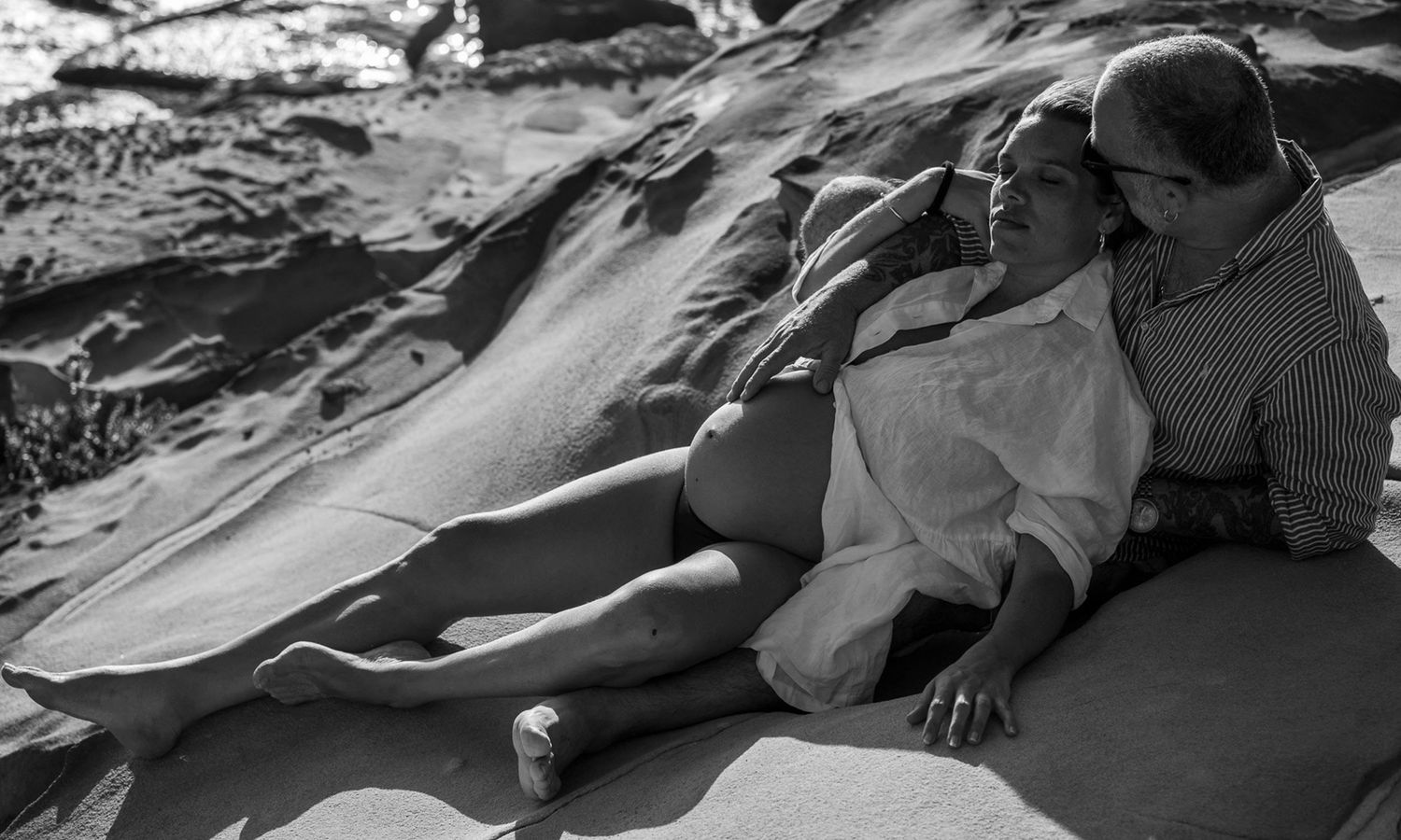 Black and white intimate outdoor photo of couple embracing on the rocks near the shoreline. Photo by Raymond Mather.