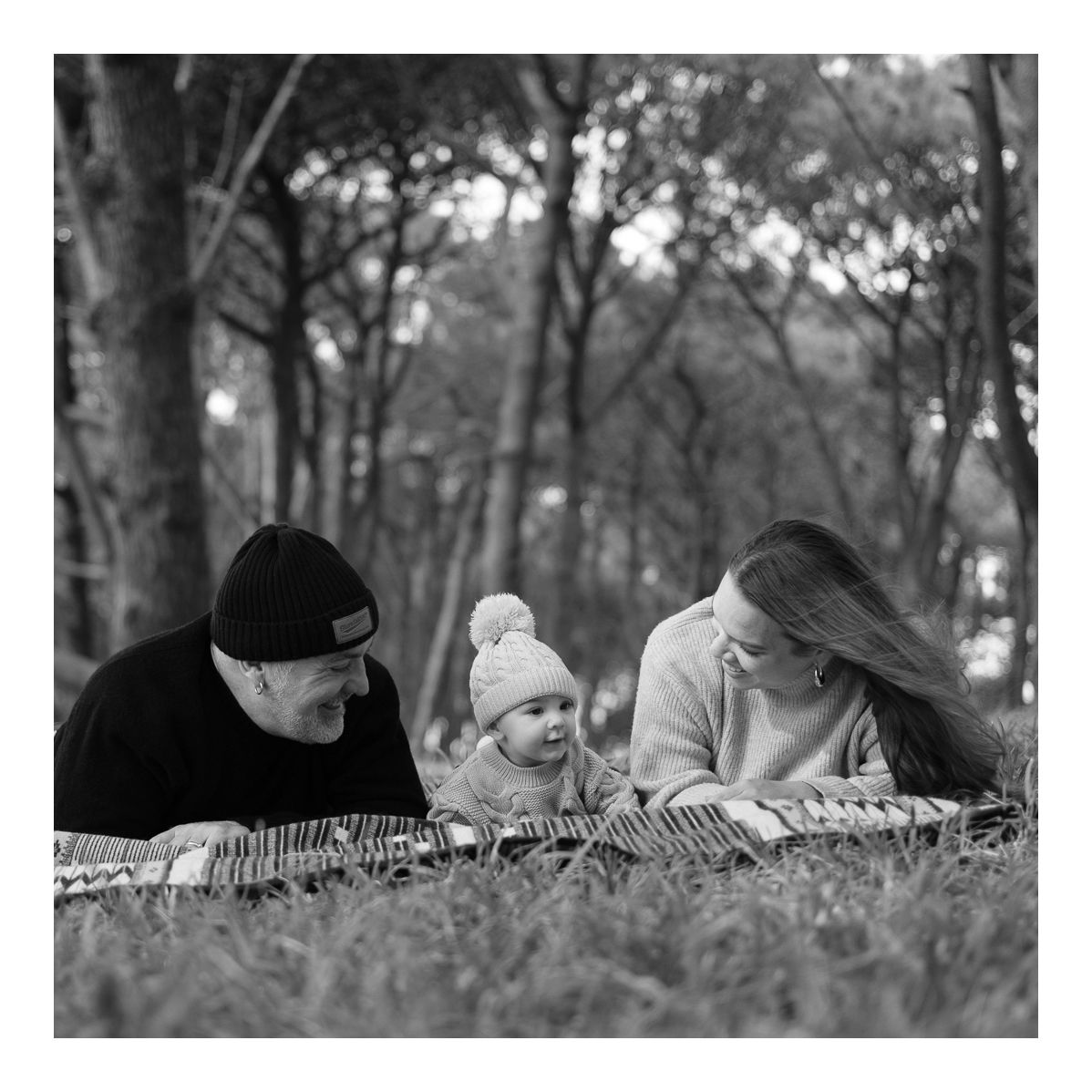 Monochrome candid photo of family lying together on a rug, sharing gentle laughter. Photo by Raymond Mather.