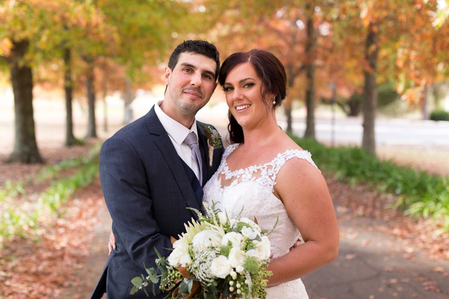 Colour couple portrait of bride and groom standing close beneath golden autumn leaves, radiating warmth and joy. Photo by Raymond Mather.