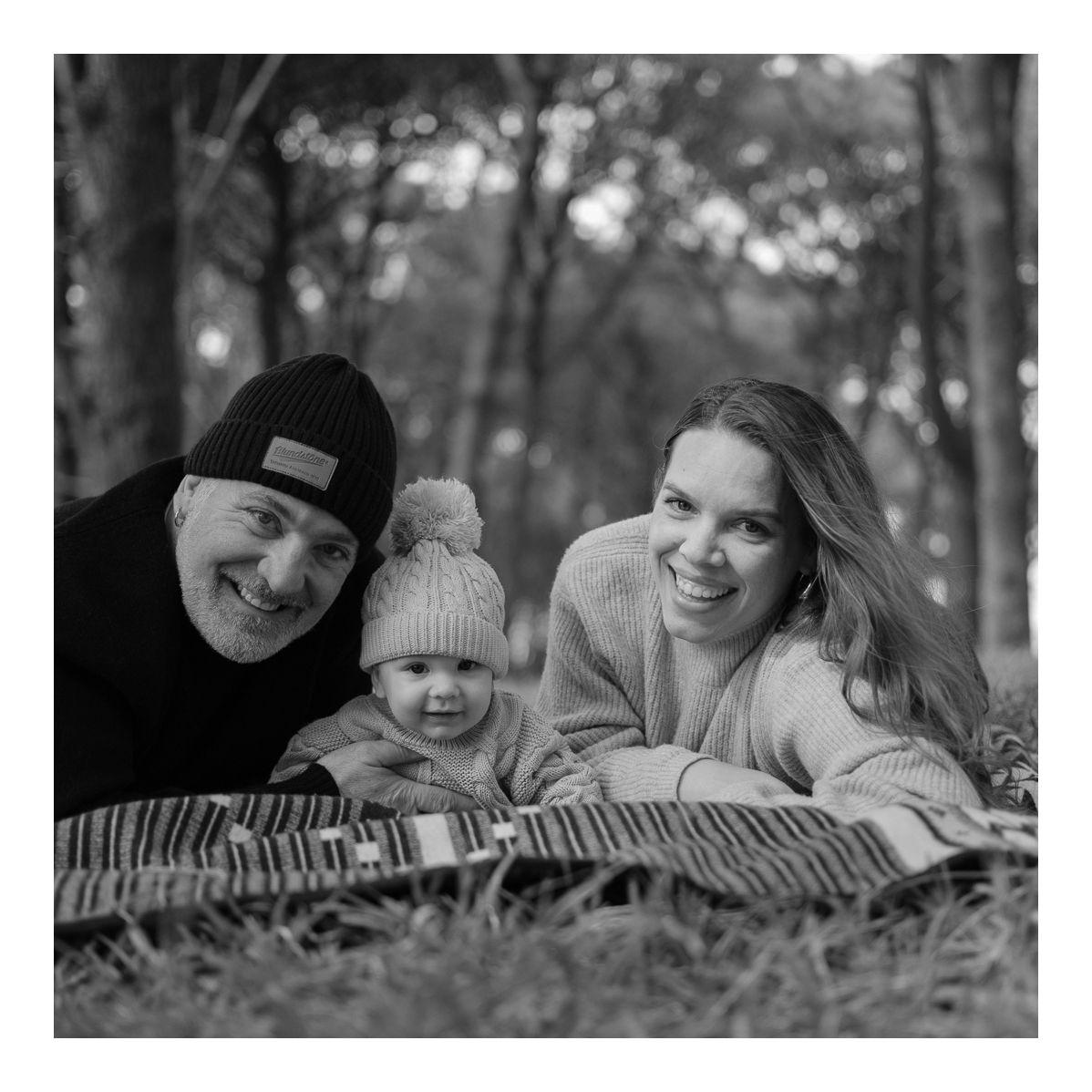 Monochrome family portrait of parents and baby lying on the grass, smiling at camera. Photo by Raymond Mather.