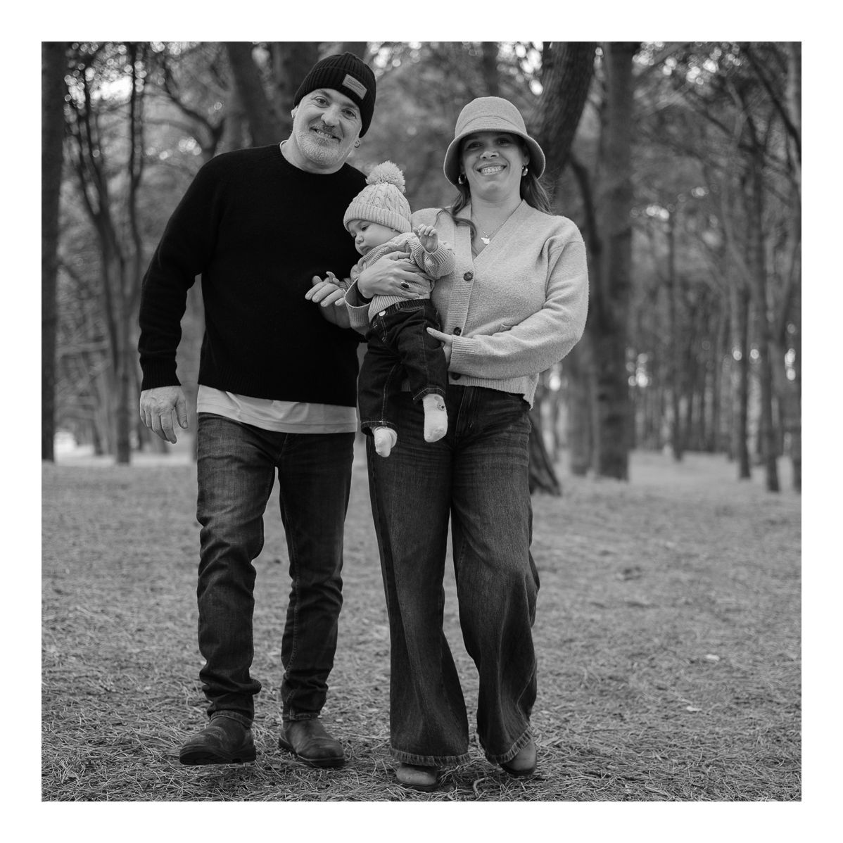 Monochrome outdoor portrait of parents walking together holding baby in the park. Photo by Raymond Mather.