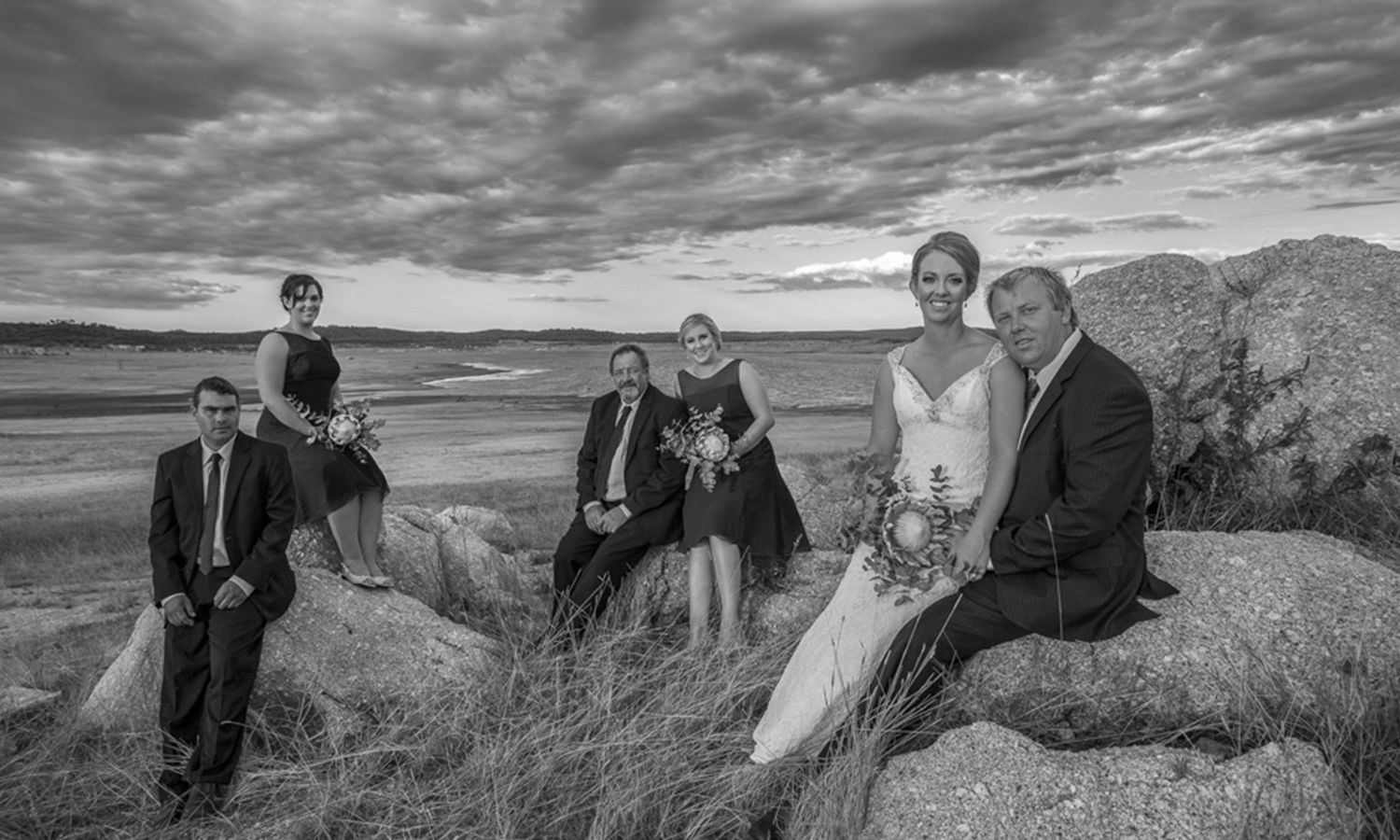 Black and white image of the wedding group seated together on shoreline rocks with scenic horizon. Photo by Raymond Mather.
