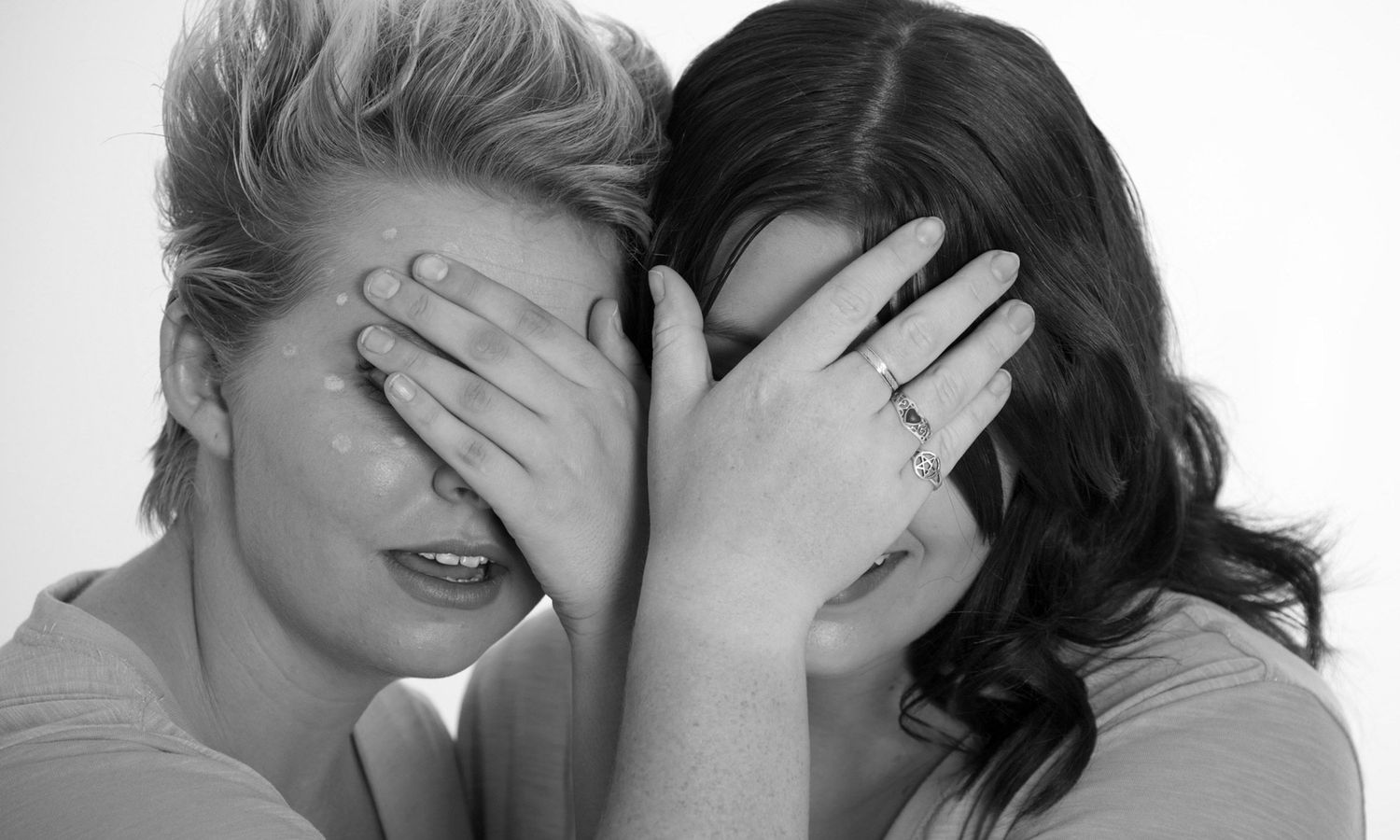 Monochrome portrait of two women laughing playfully while covering each other’s eyes. Photo by Raymond Mather.