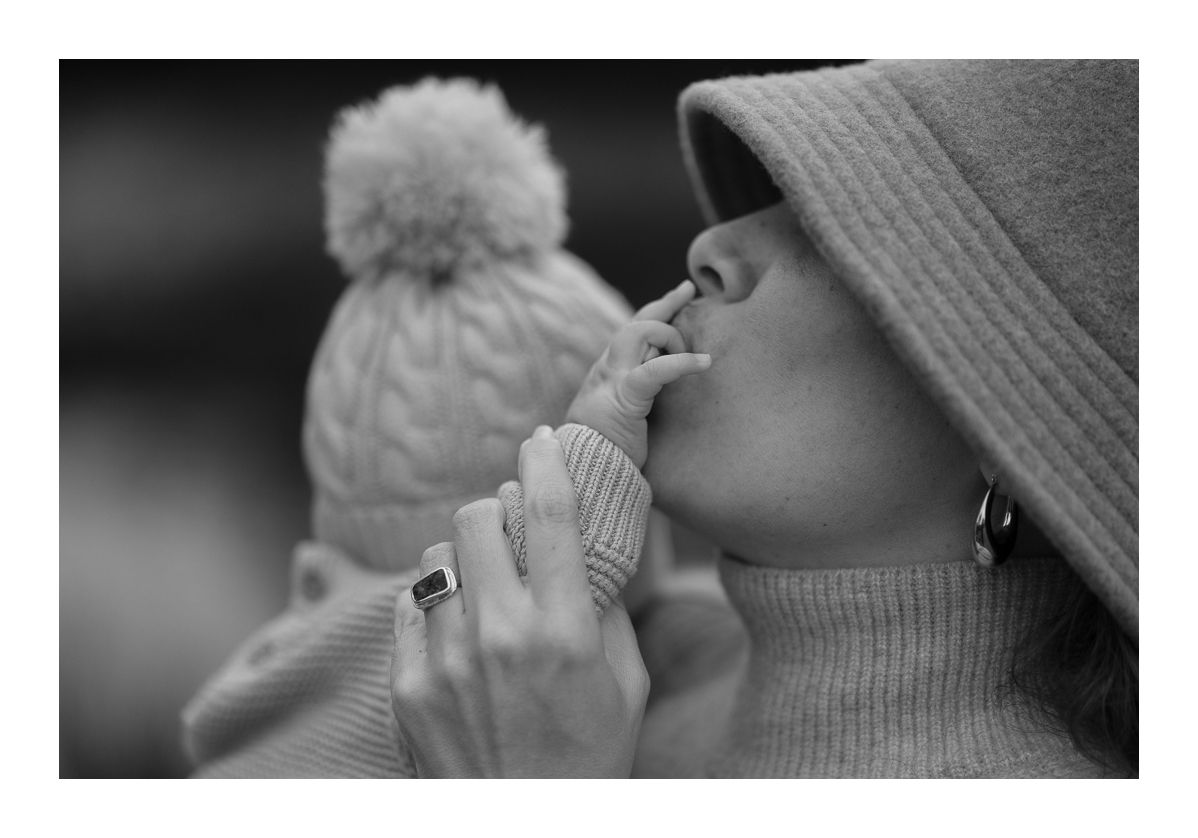 Monochrome close-up outdoor photo of mother kissing baby’s hand in winter outfits. Photo by Raymond Mather.
