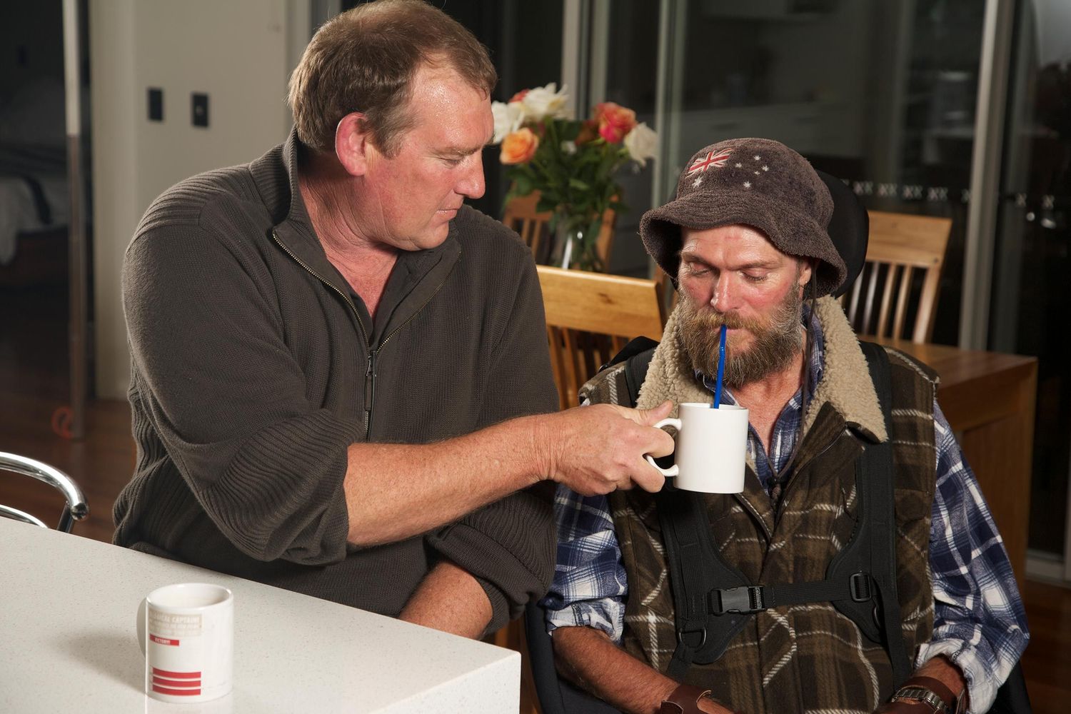 Two men at kitchen table, one assisting the other with a warm drink, a candid expression of empathy, everyday care. Photo by Raymond Mather.