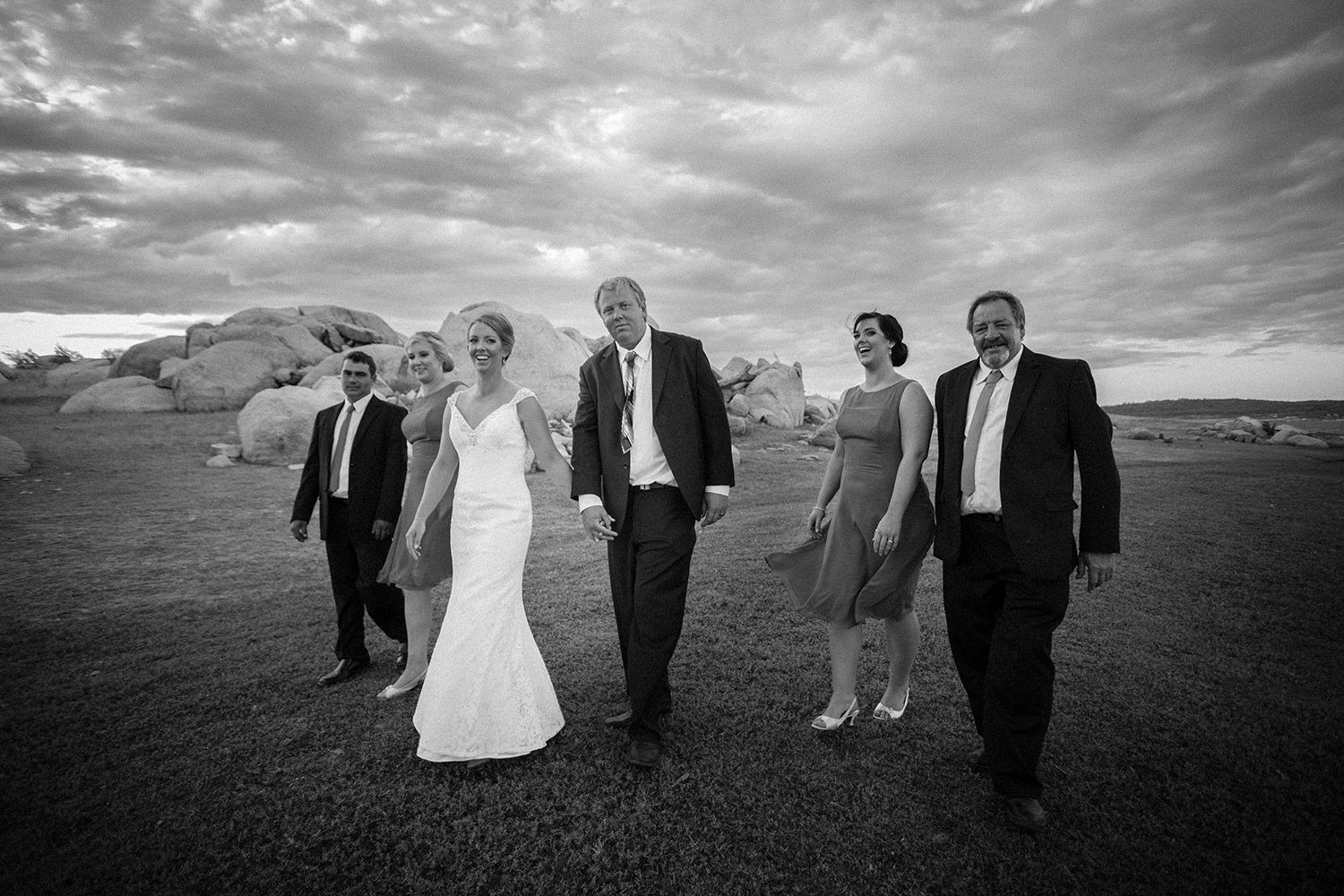 Monochrome wedding party photo of couple walking joyfully with friends along a windswept lake. Photo by Raymond Mather.