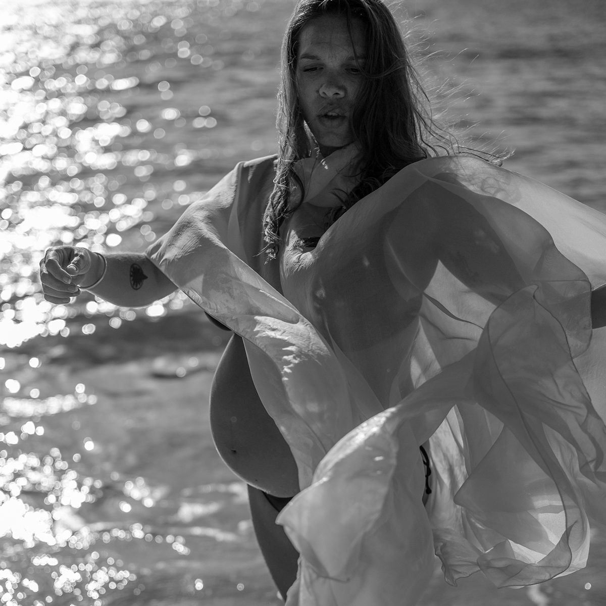 Monochrome artistic maternity portrait of woman standing in ocean light with flowing fabric. Photo by Raymond Mather.