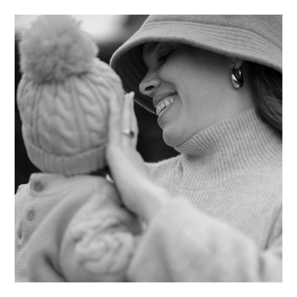 Black and white candid outdoor photo of mother smiling at her baby in winter clothes. Photo by Raymond Mather.