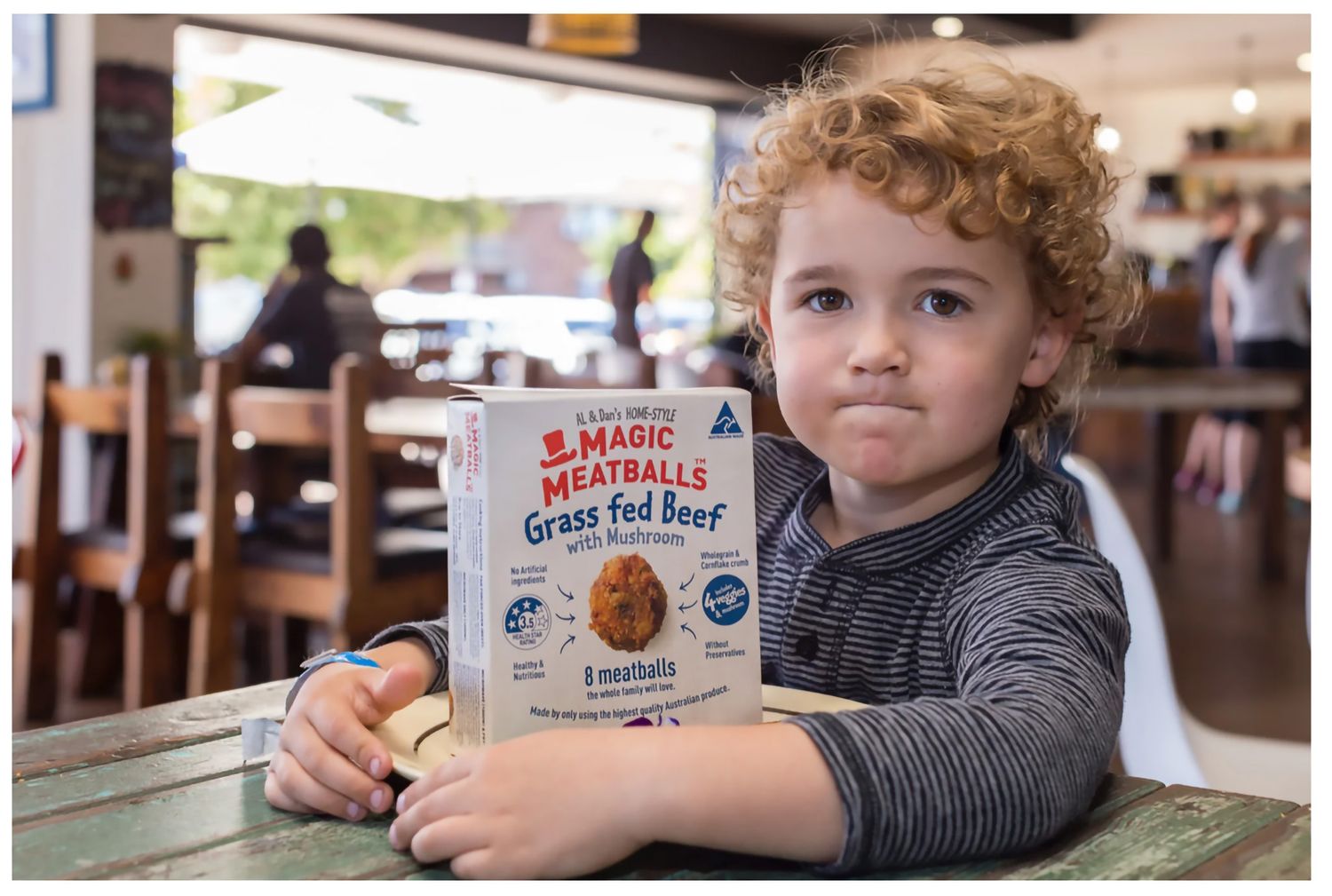 Young curly-haired child holding Magic Meatballs Grass Fed Beef box at a restaurant table.