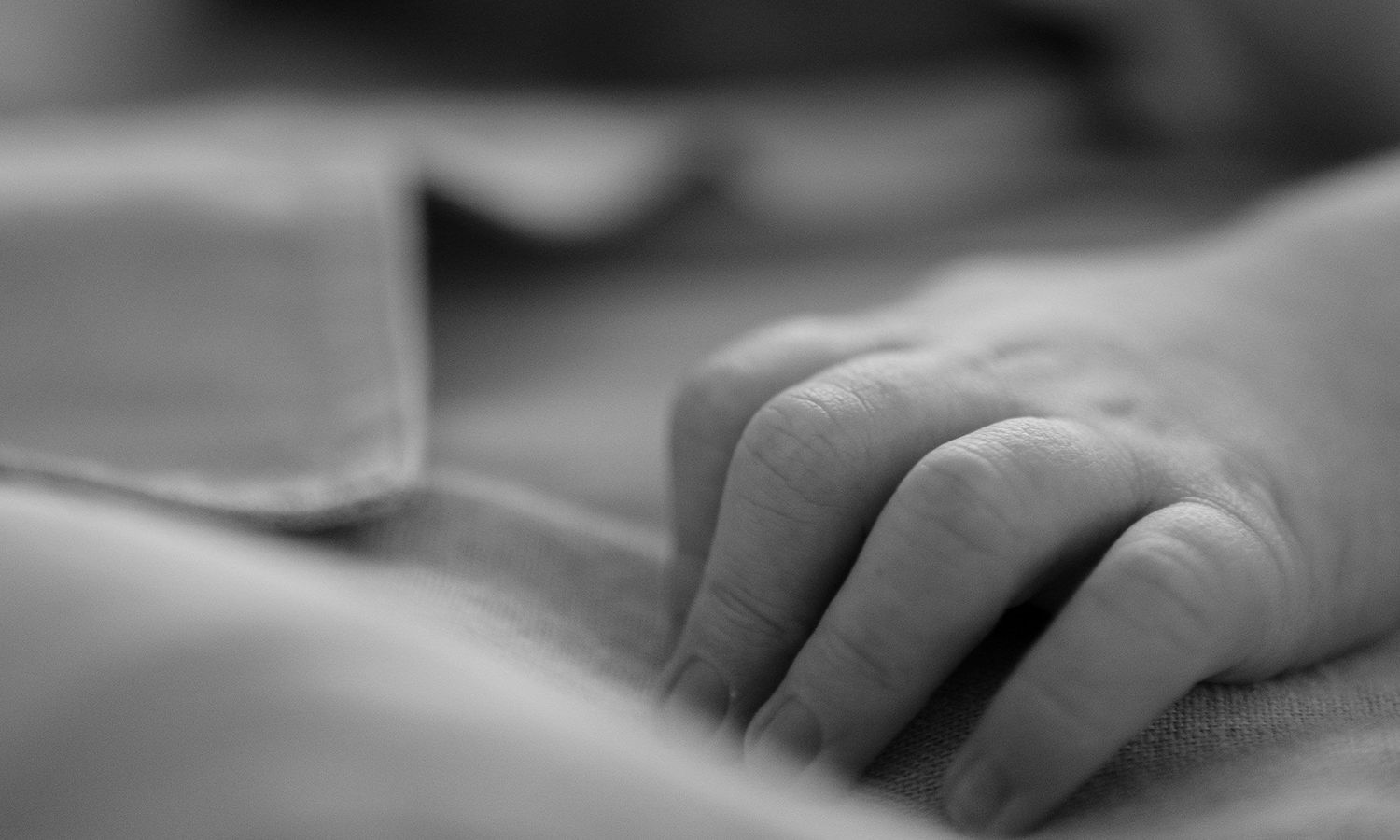 Monochrome close-up photo of baby’s hand resting gently on parent’s arm. Photo by Raymond Mather.