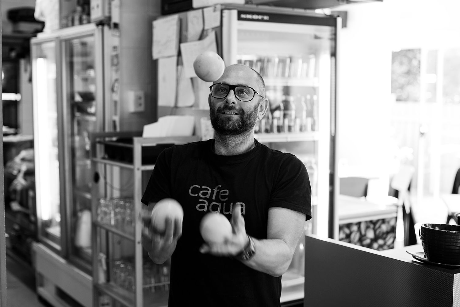 Black and white lifestyle portrait of a barista juggling oranges playfully at Café. Photo by Raymond Mather.