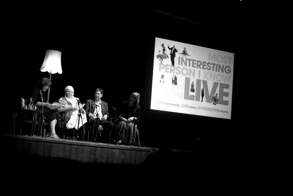 Black and white photo of panelists on stage at Most Interesting Person I Know Live event with projected screen.