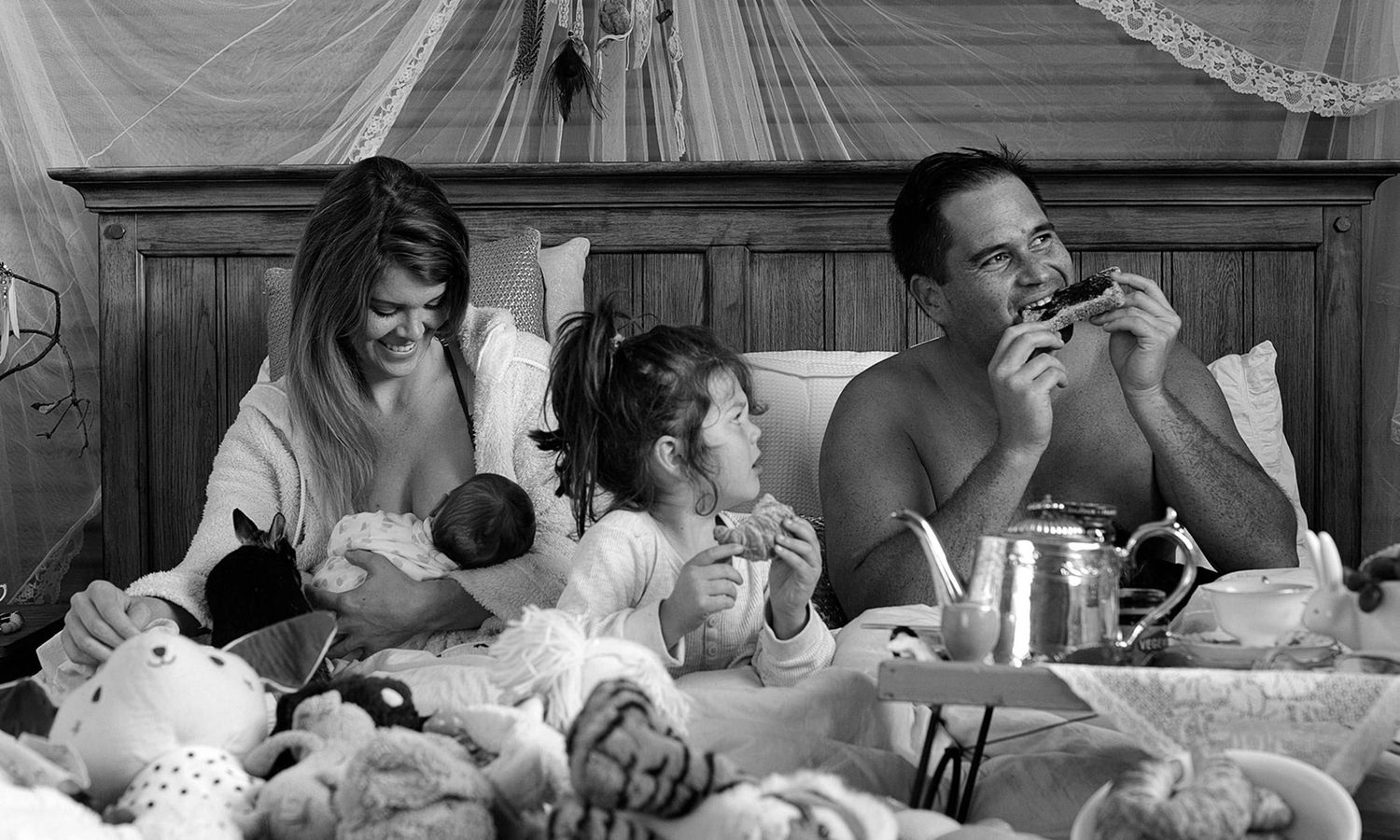 Black and white candid photo of family enjoying breakfast in bed, full of laughter and warmth. Photo by Raymond Mather.