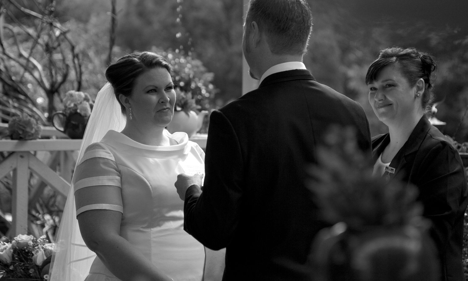 Black and white wedding photo of bride and groom exchanging vows beneath soft garden light. Photo by Raymond Mather.