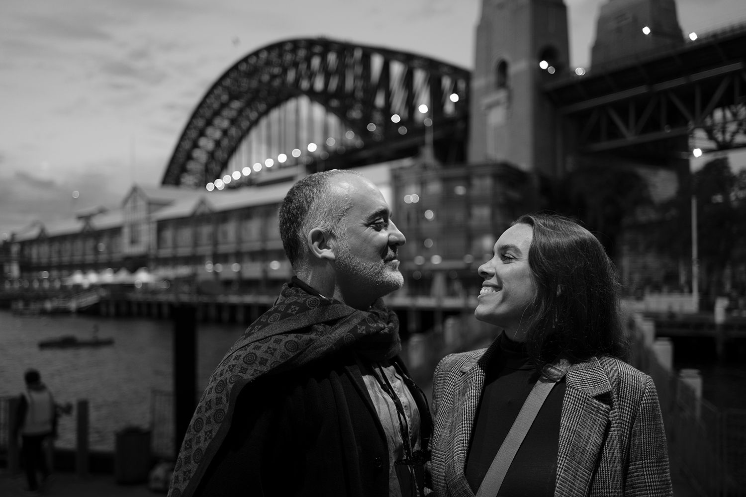 Black and white evening portrait of a couple smiling beneath Sydney Harbour Bridge. Photo by Raymond Mather.