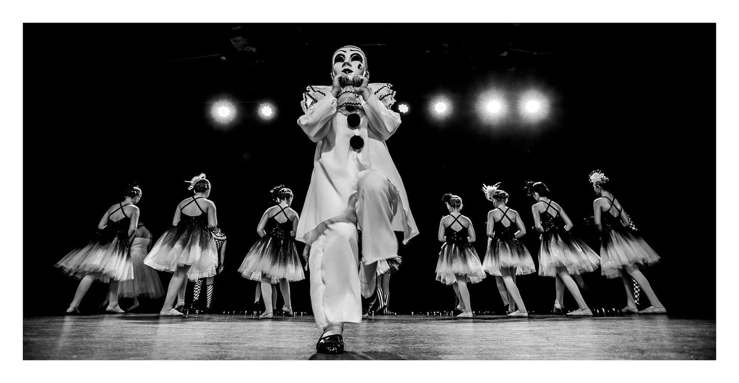Black and white photo of a mime clown performing on stage with ballet dancers in tutus lined up behind them.