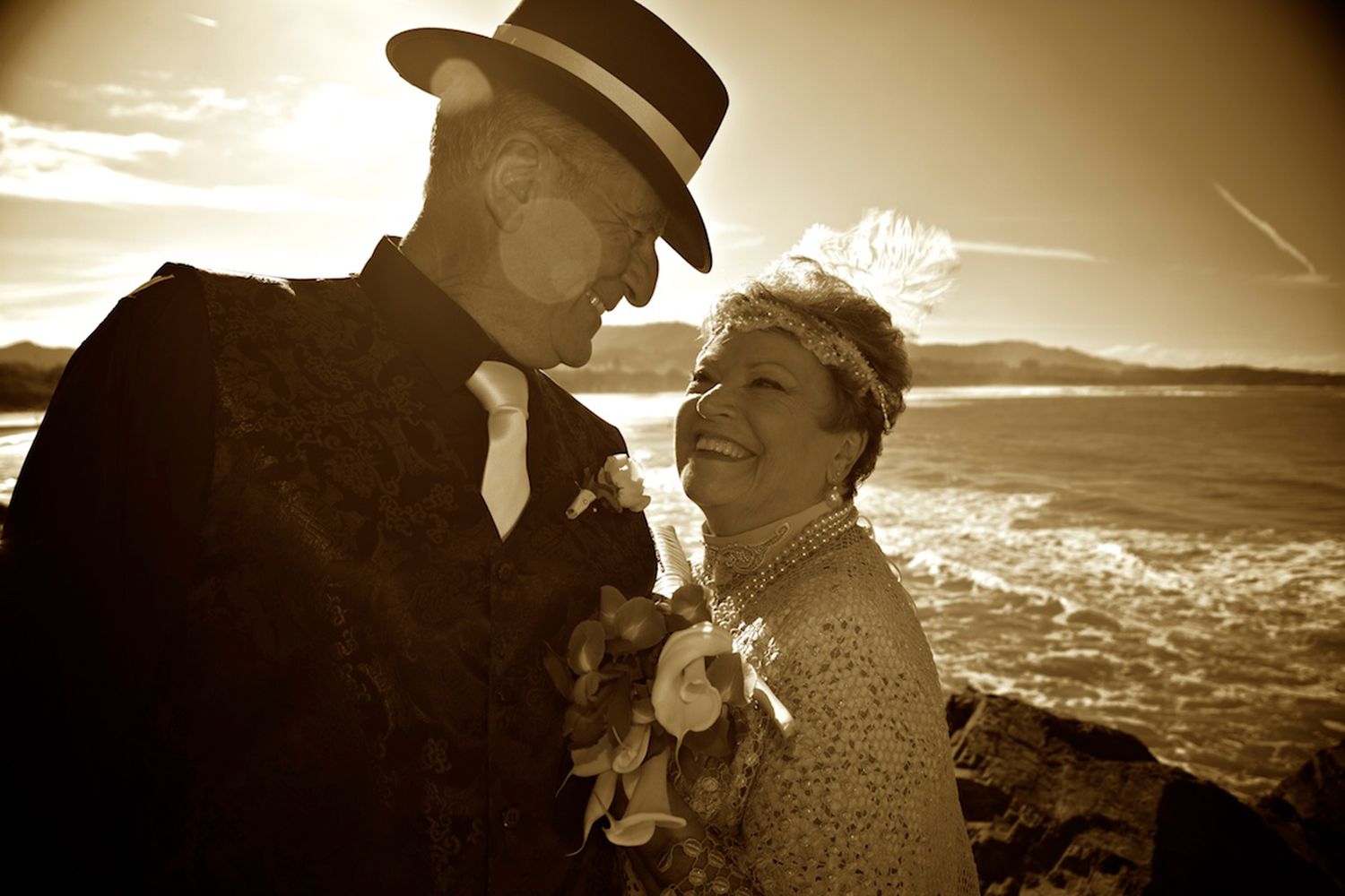 Monochrome sepia wedding photo of couple smiling warmly against golden light and ocean backdrop. Photo by Raymond Mather.