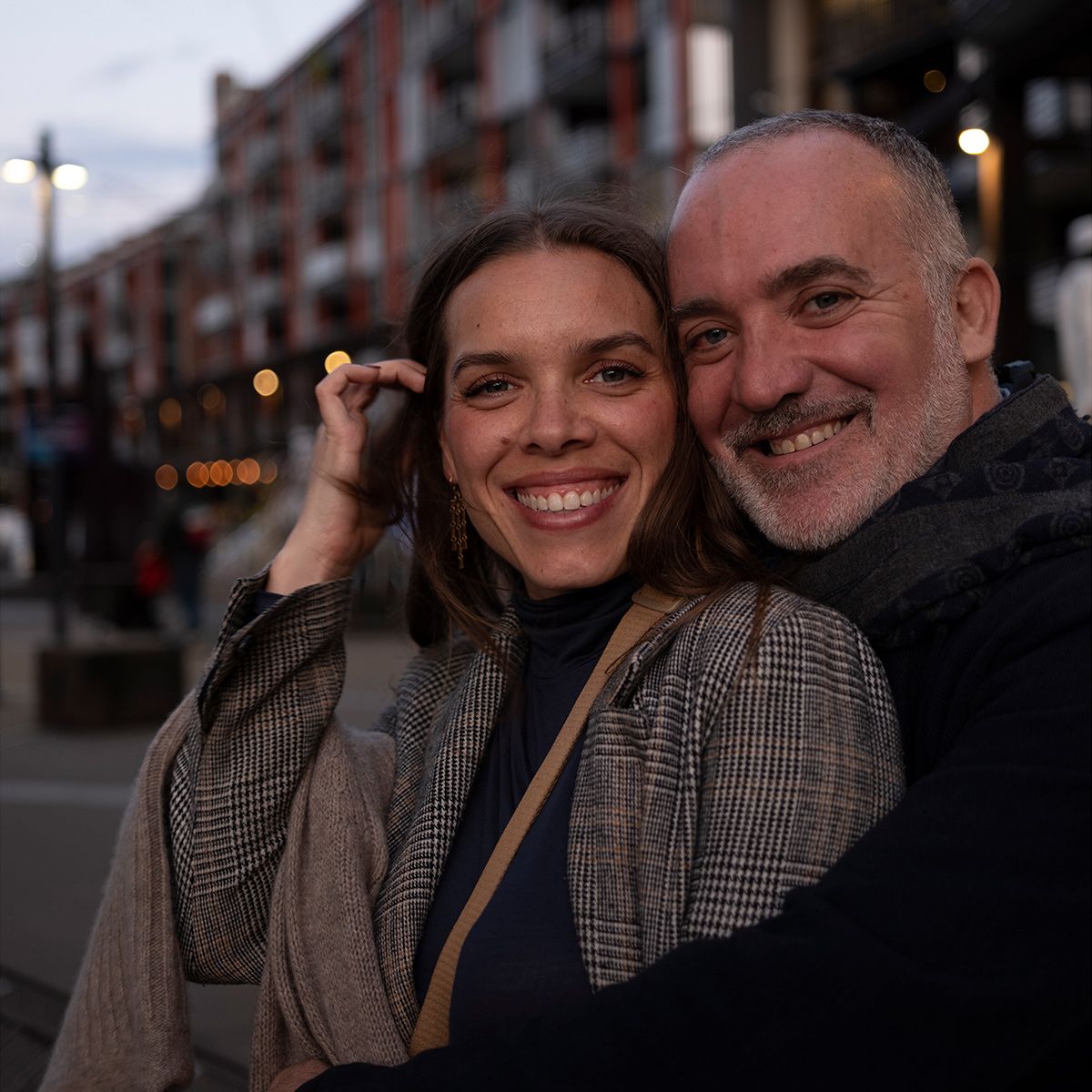 A couple shares a tender moment together on a city street at dusk, with buildings and streetlights in the background.