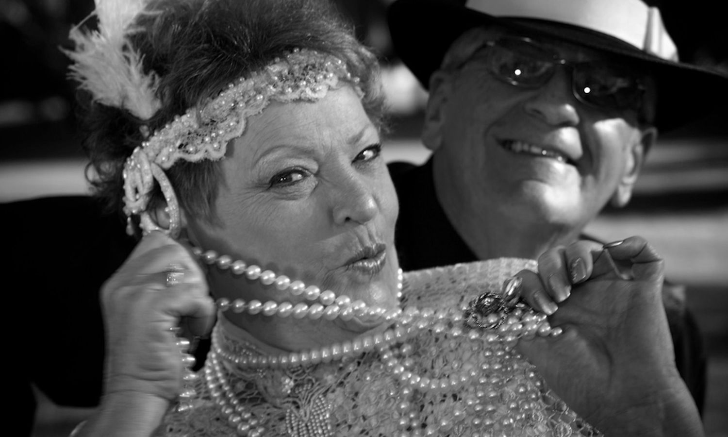 Black and white vintage-style photo of smiling couple dressed in 1920s fashion, framed with playful elegance. Photo by Raymond Mather.