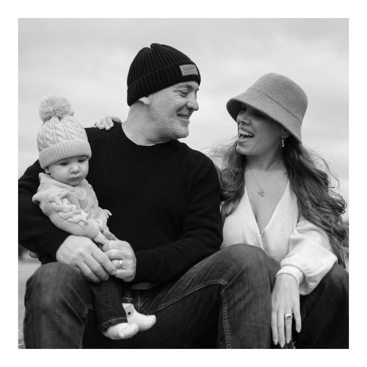 Monochrome outdoor portrait of family sitting together, smiling warmly on a cool day. Photo by Raymond Mather.
