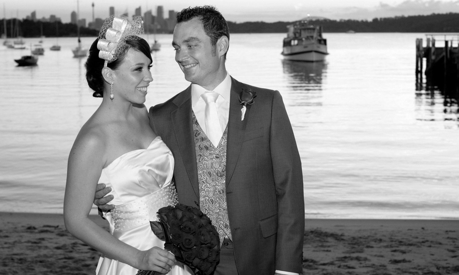 Black and white portrait of bride and groom standing by the harbour at sunset, framed against the skyline. Photo by Raymond Mather.